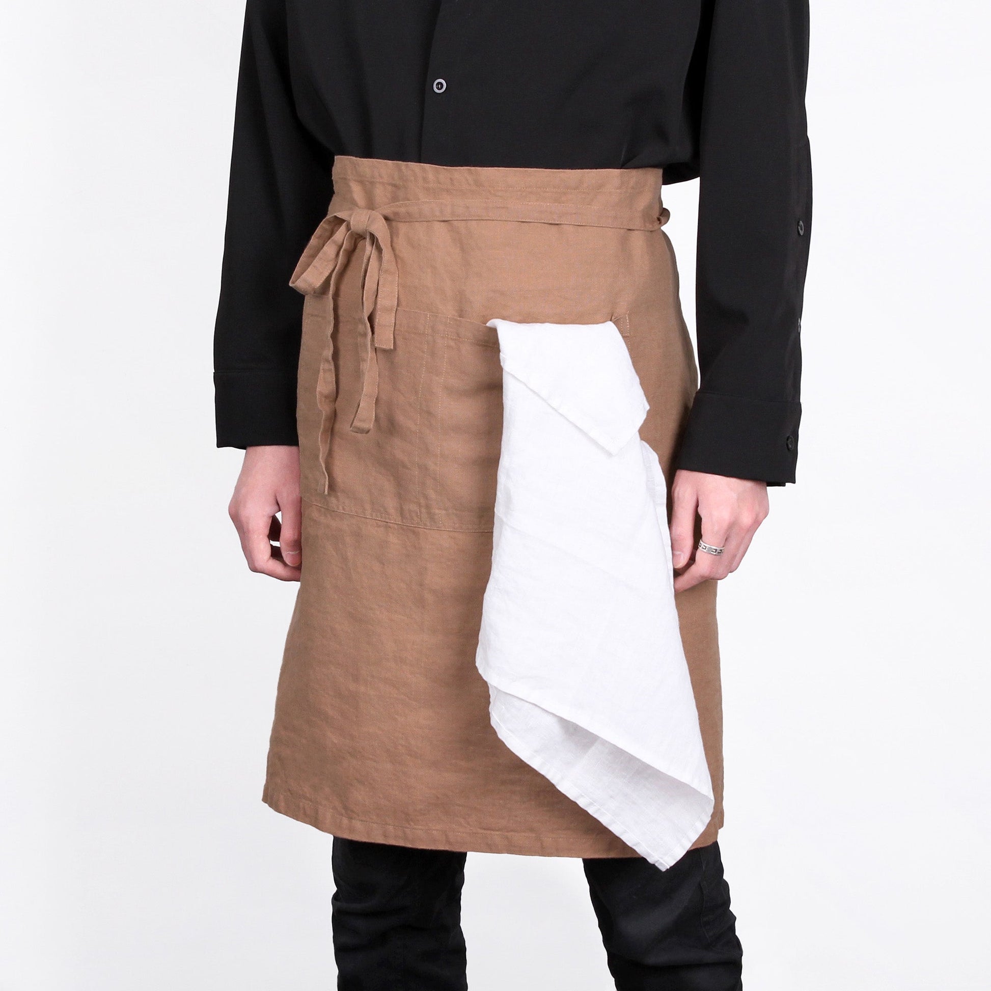 A person wearing a brown linen-style apron with a white cloth tucked under one side, paired with a black long-sleeve shirt and black pants, against a plain white background.