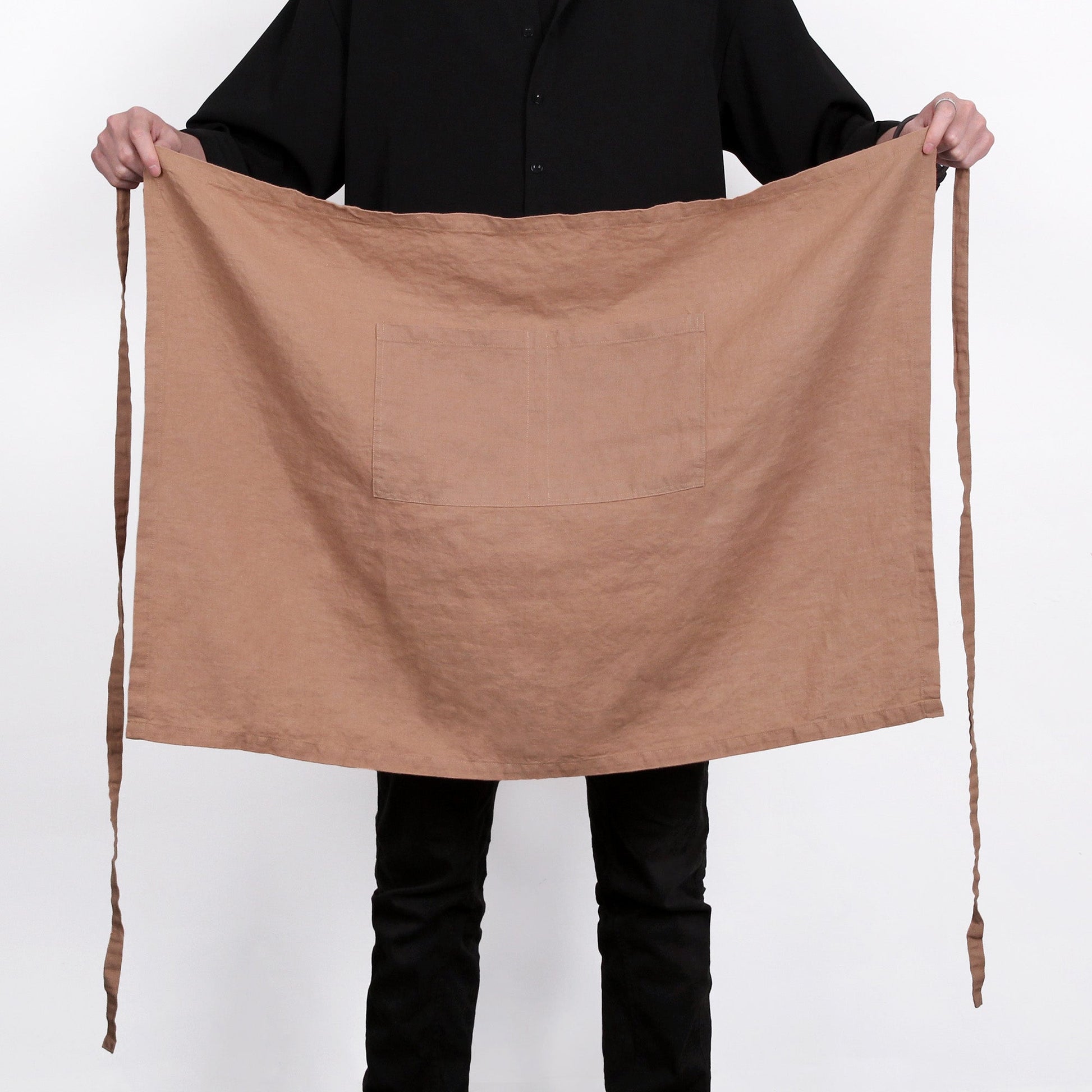 A person wearing a black shirt and black pants holds up a large, plain brown linen apron with a single front pocket and long ties, set against a plain white background.