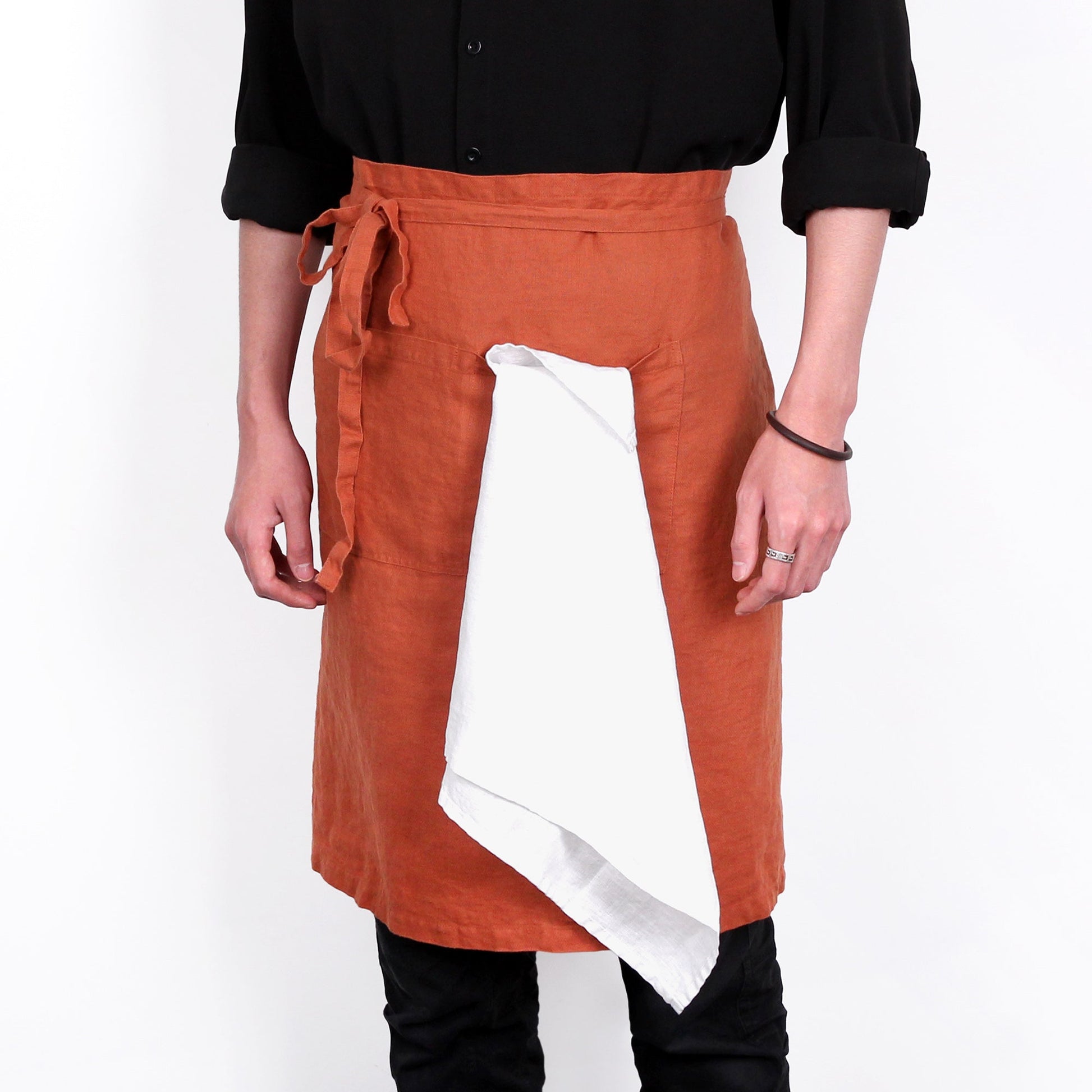 A person wearing a rust-orange wrap-style apron with a white inner panel, paired with a black shirt and black pants, shown against a plain white background.