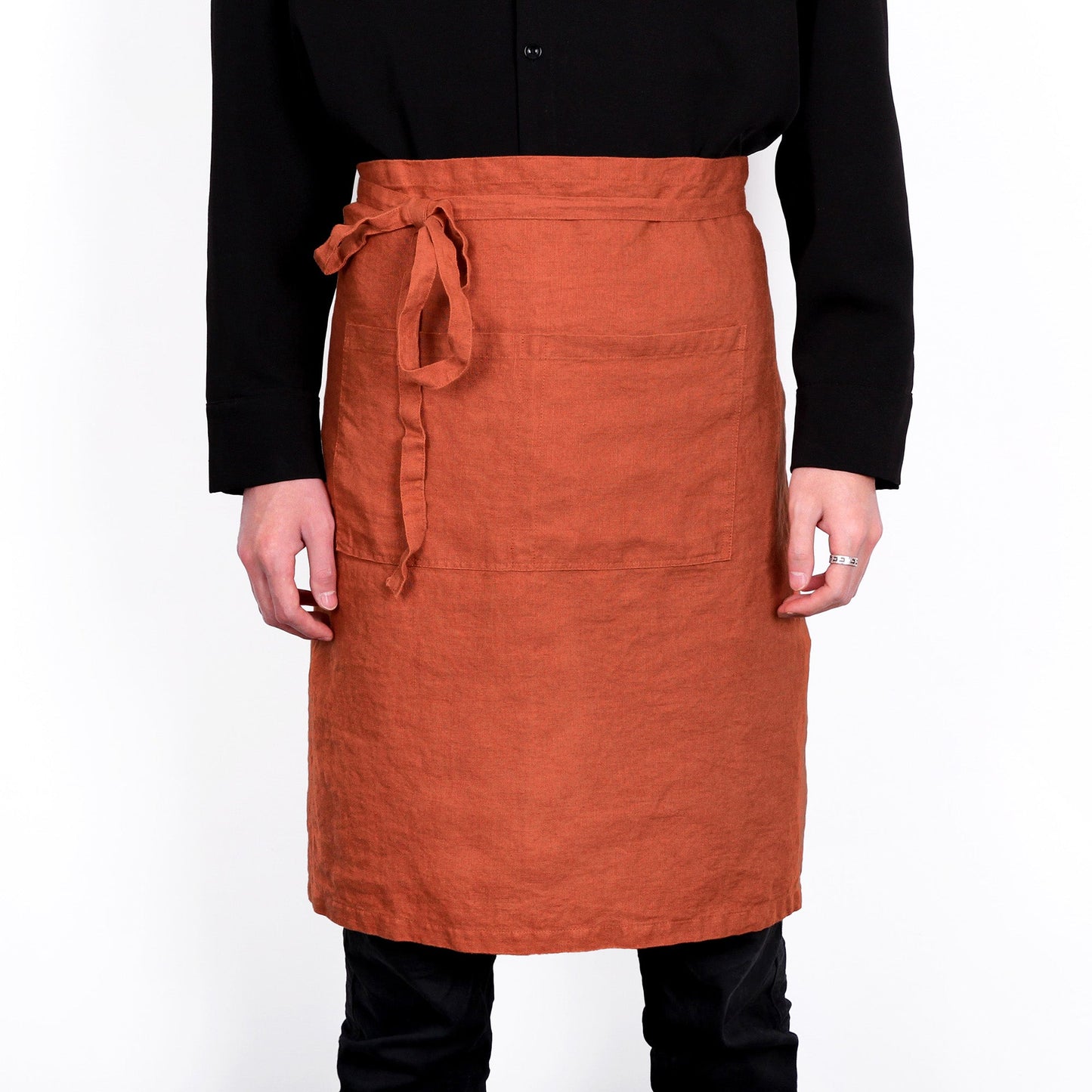 A terracotta orange linen bib apron with a matching waist tie, worn by a model in a black long-sleeve shirt and black pants against a white background.