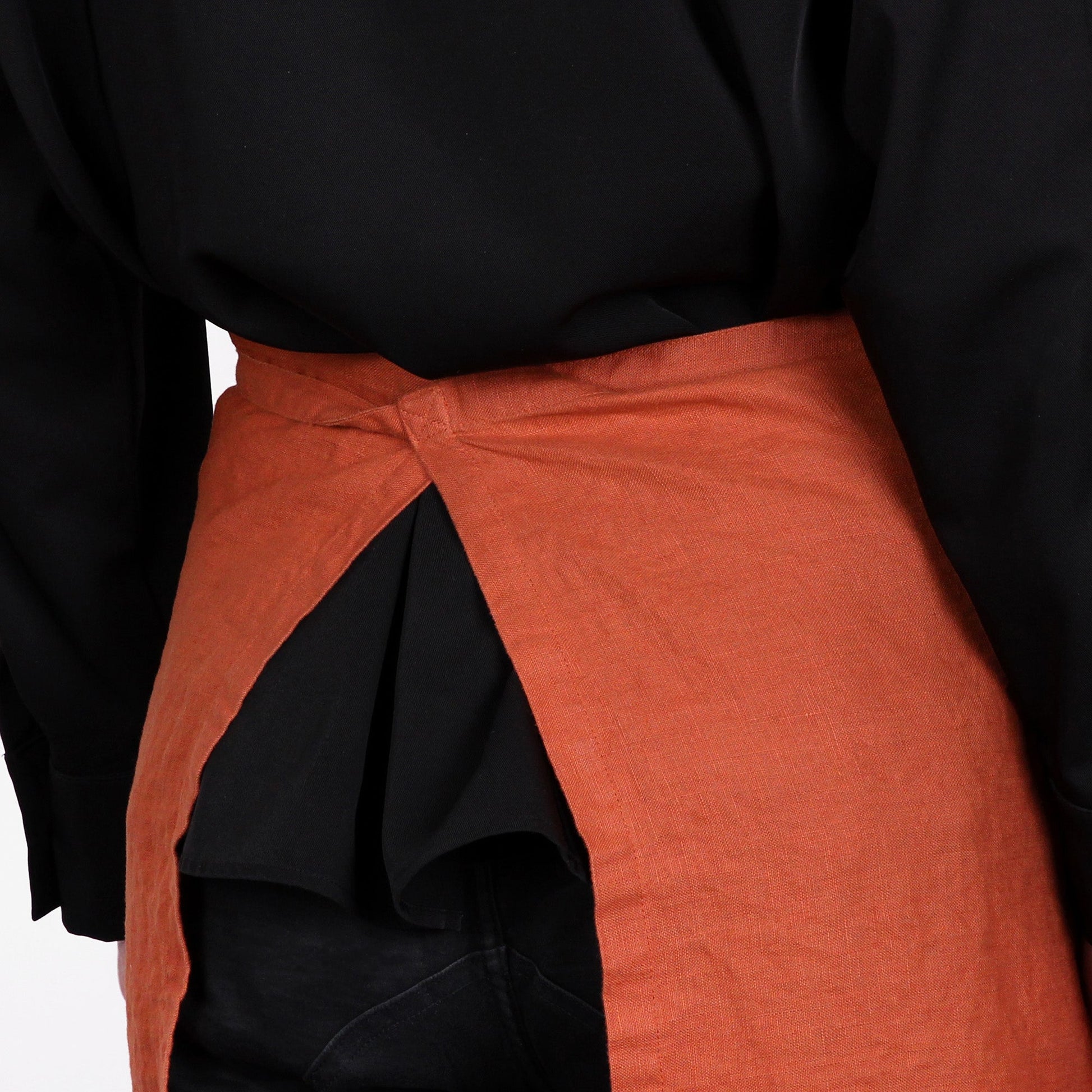 A close-up of a person wearing a rust-orange linen apron tied around the waist over a black top and black pants.