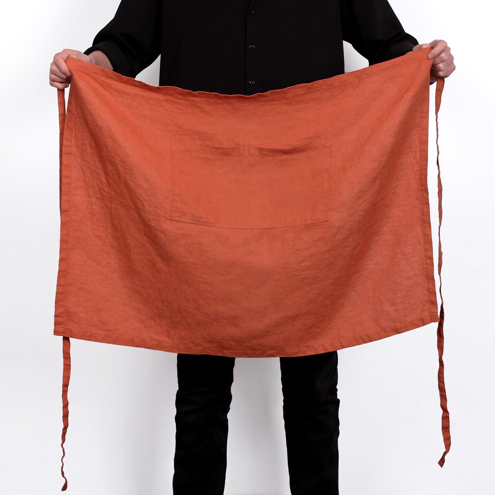A person wearing a black shirt and black pants holds up a large terracotta orange linen apron with a front pocket and tie strings against a white background.