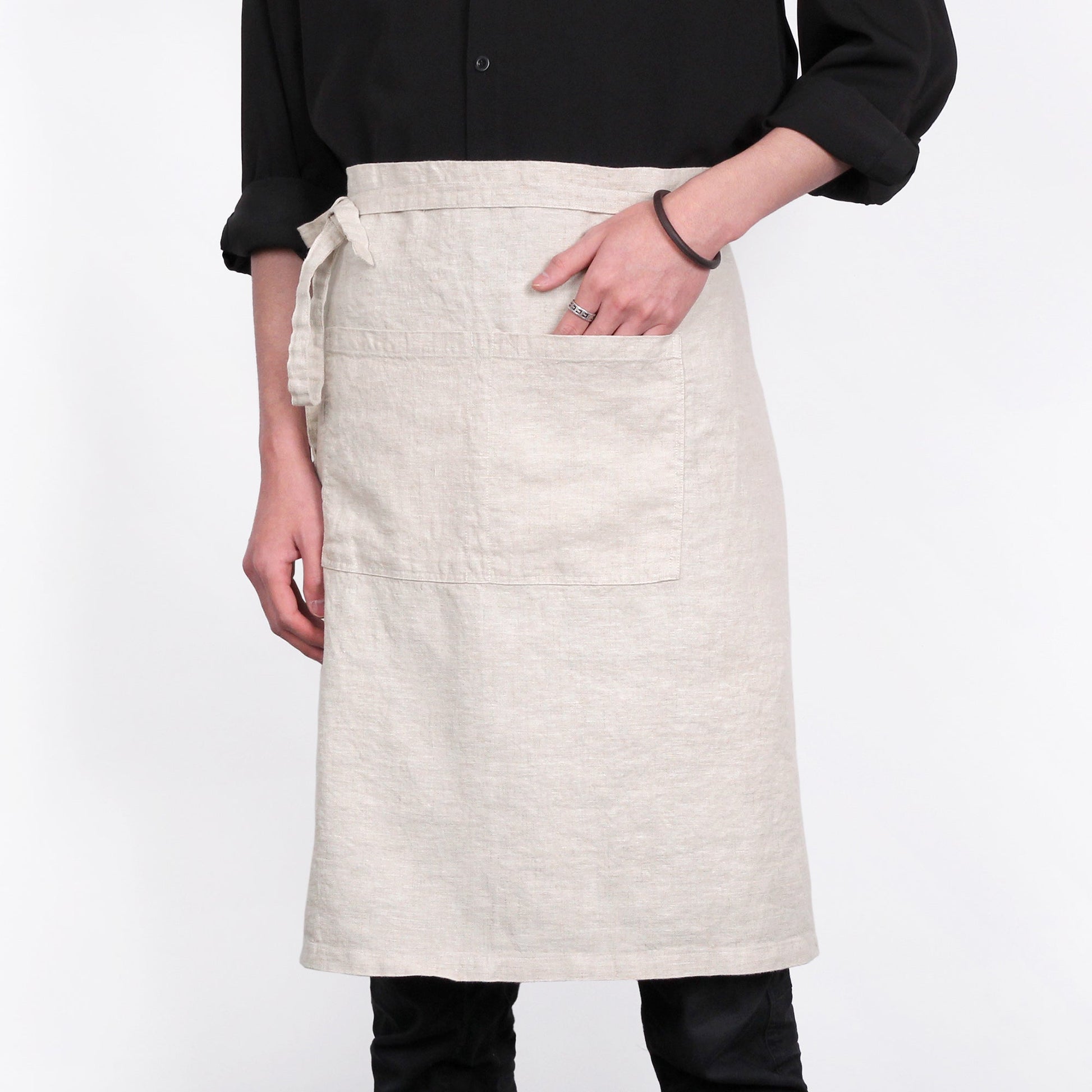 A person wearing a light beige linen apron with a front pocket over a black shirt, shown from the chest down against a plain white background.
