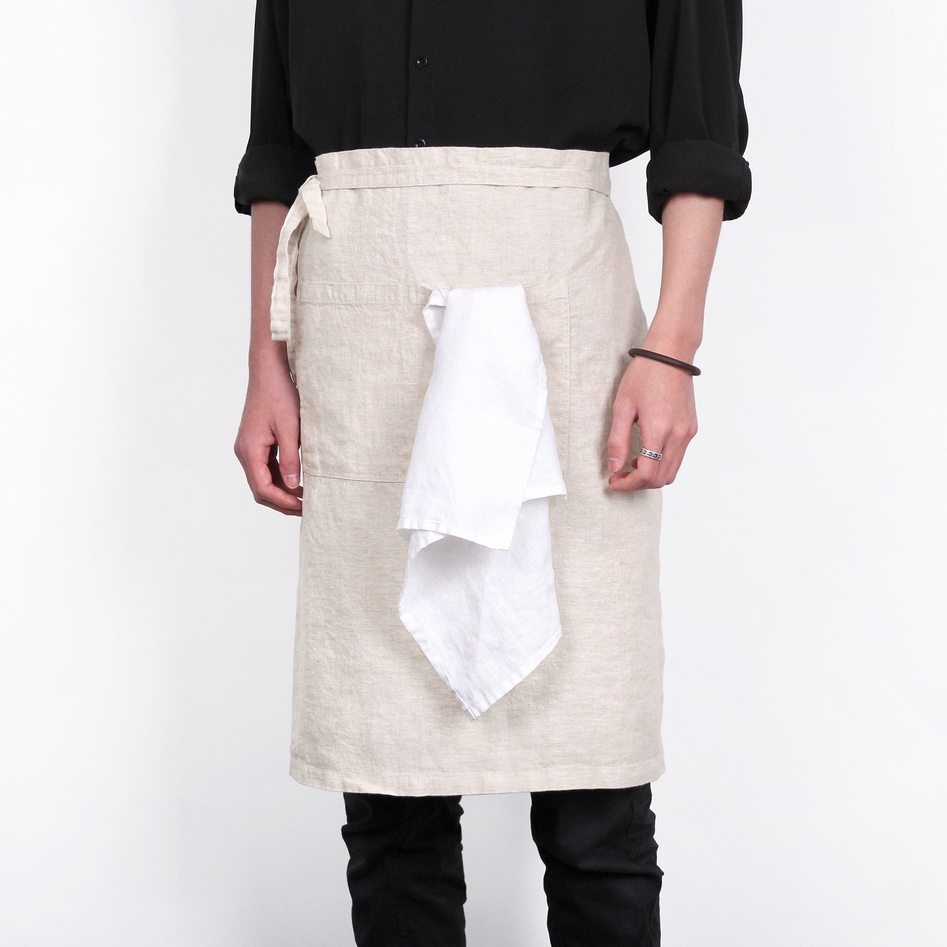 A person wearing a light beige linen apron with a white cloth tucked into the front pocket, paired with a black shirt and black pants, against a plain white background.
