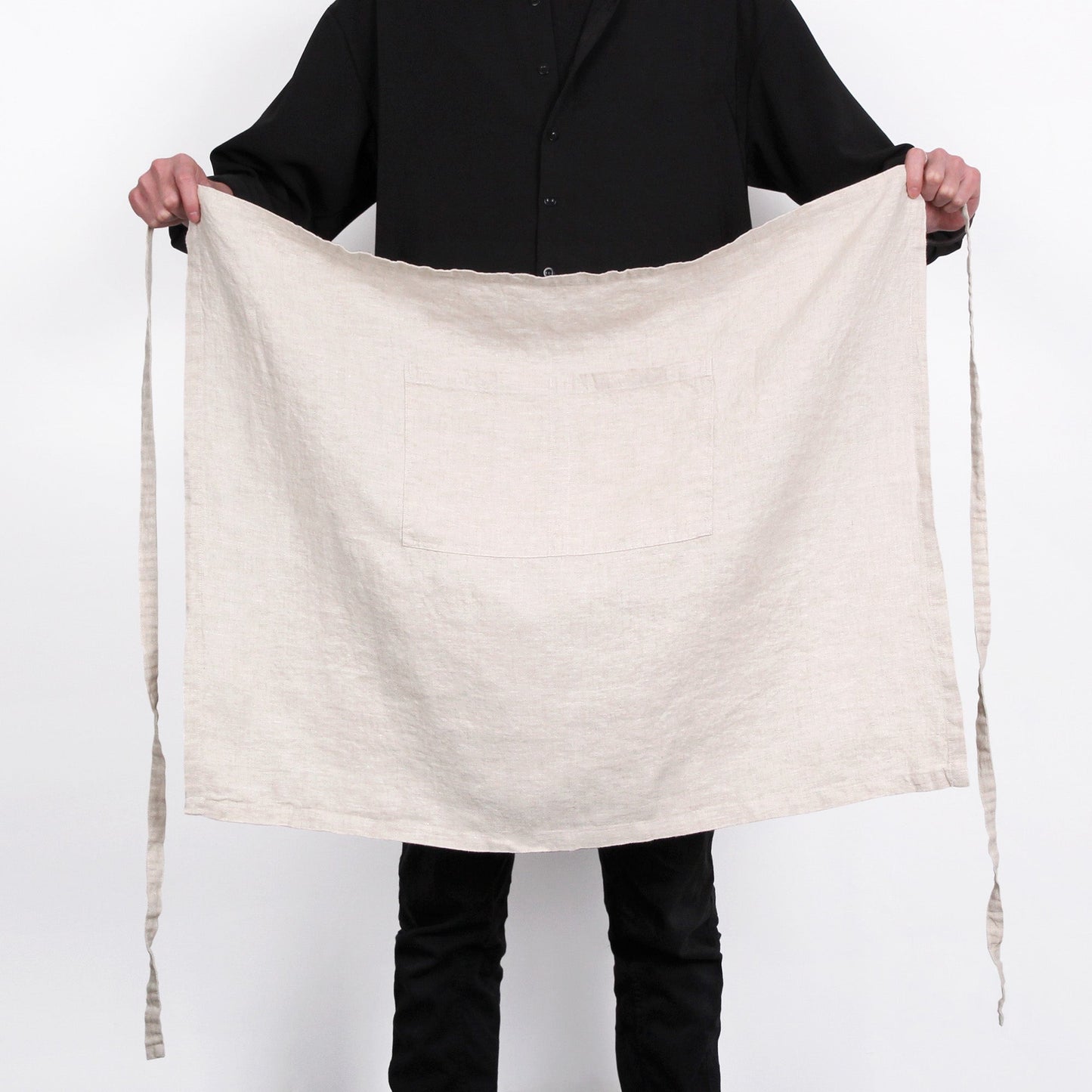 A person wearing a black shirt and black pants holds up a large, square, off-white linen apron with a single front pocket and long ties, set against a plain white background.