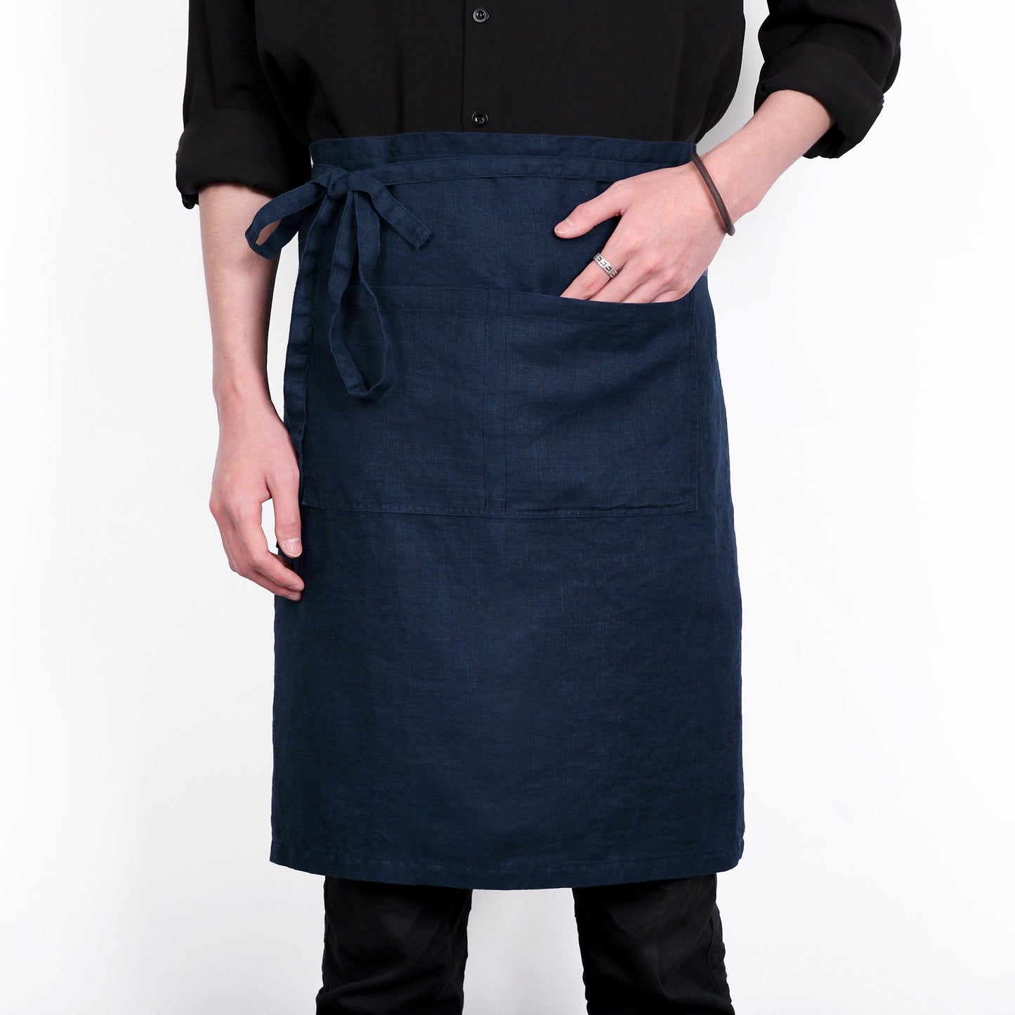 A dark navy blue linen apron with a tie waist and front pocket, worn by a model in a black shirt against a white background.