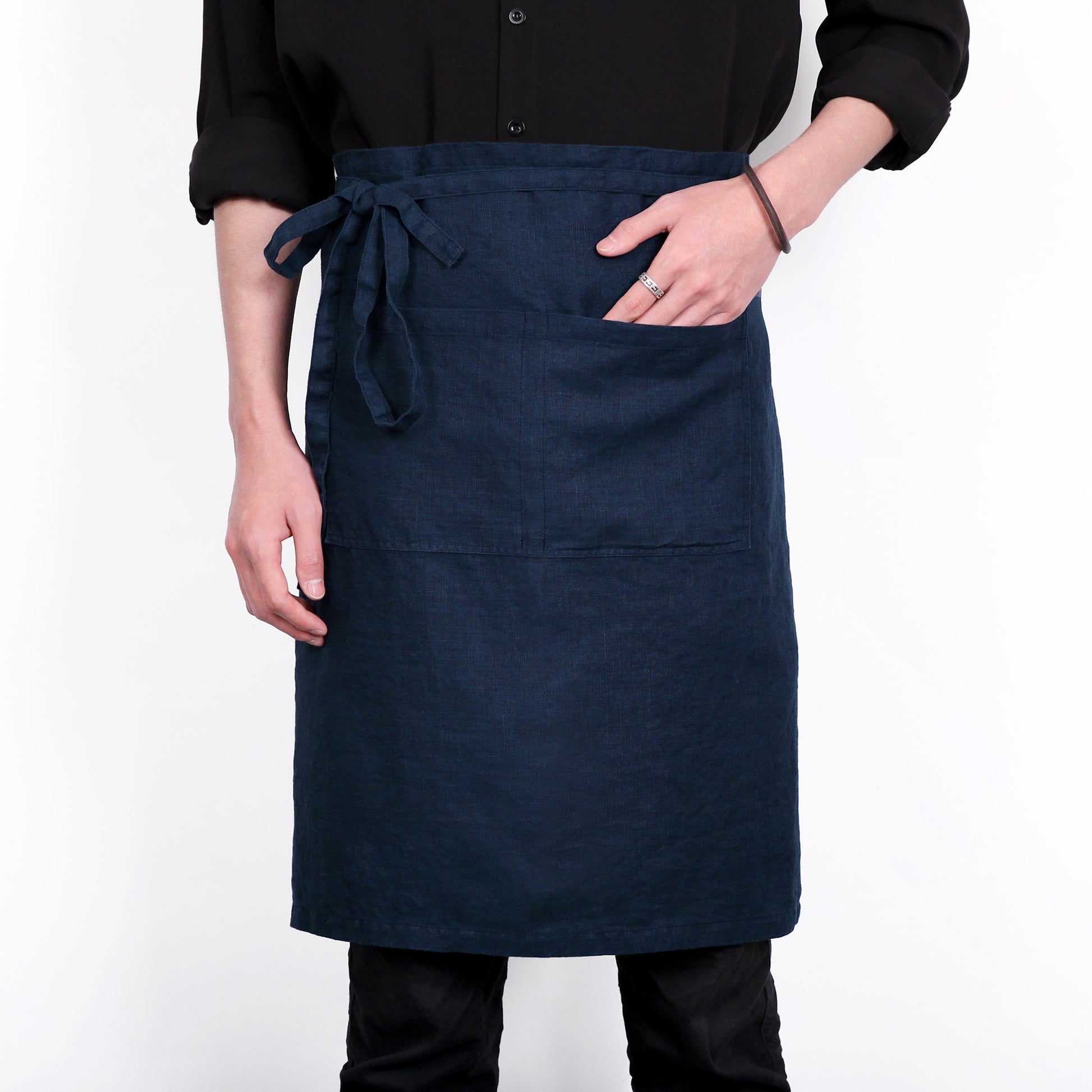 A dark navy blue linen apron with a tie waist and front pocket, worn by a model in a black shirt against a white background.