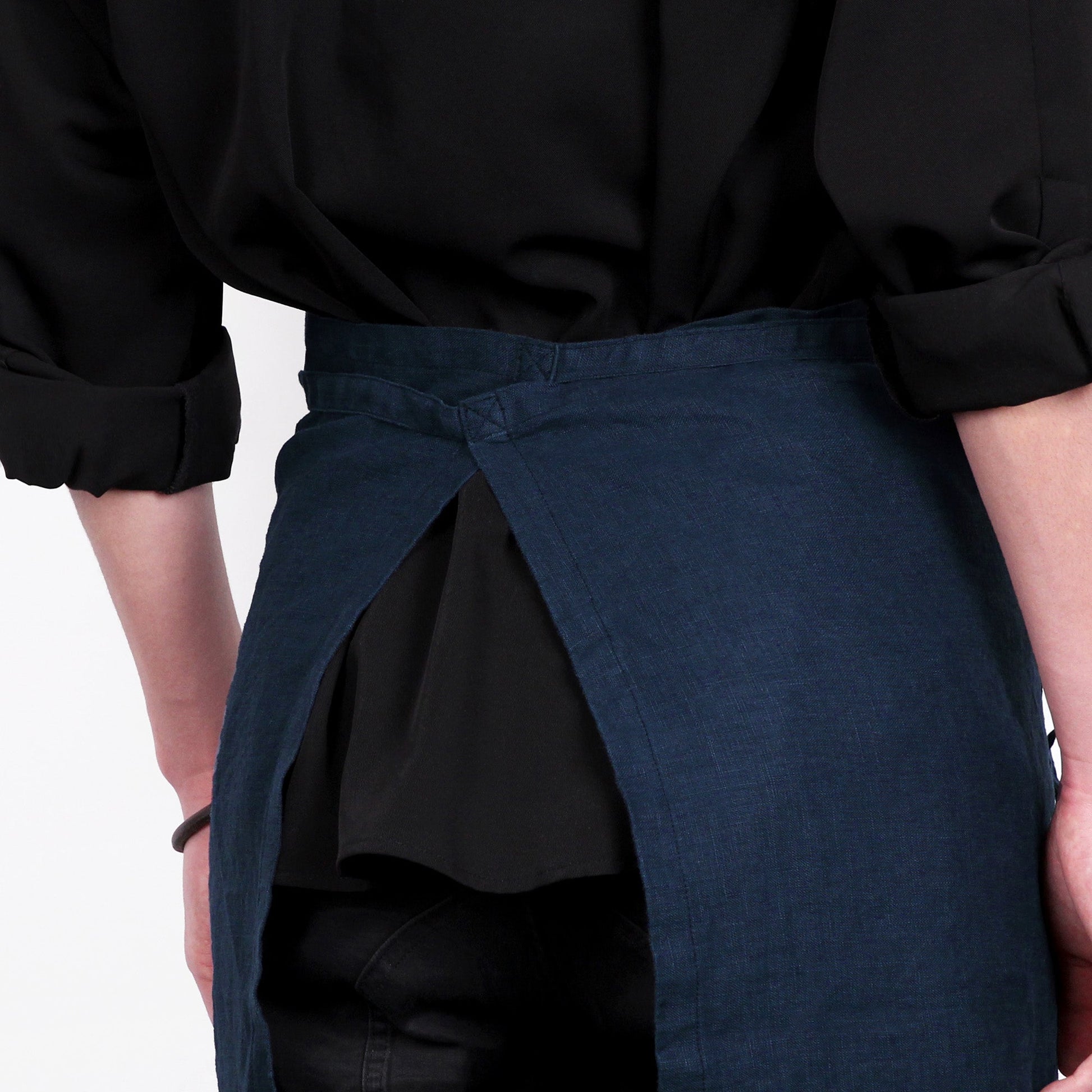 A close-up of a person wearing a dark navy wrap apron over a black shirt, showing the waist tie and front drape of the apron.