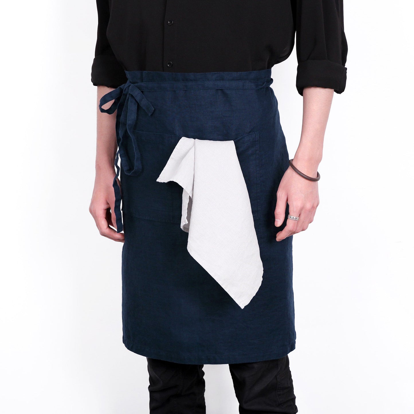 A person wearing a dark navy apron over a black shirt and black pants, with a white cloth tied around the waist, photographed against a plain white background.