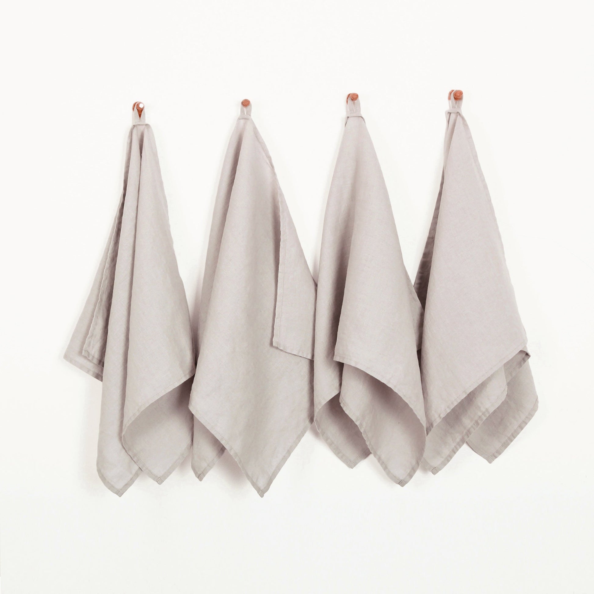 Four light taupe linen kitchen tea towels hanging from small wooden pegs on a plain white wall.