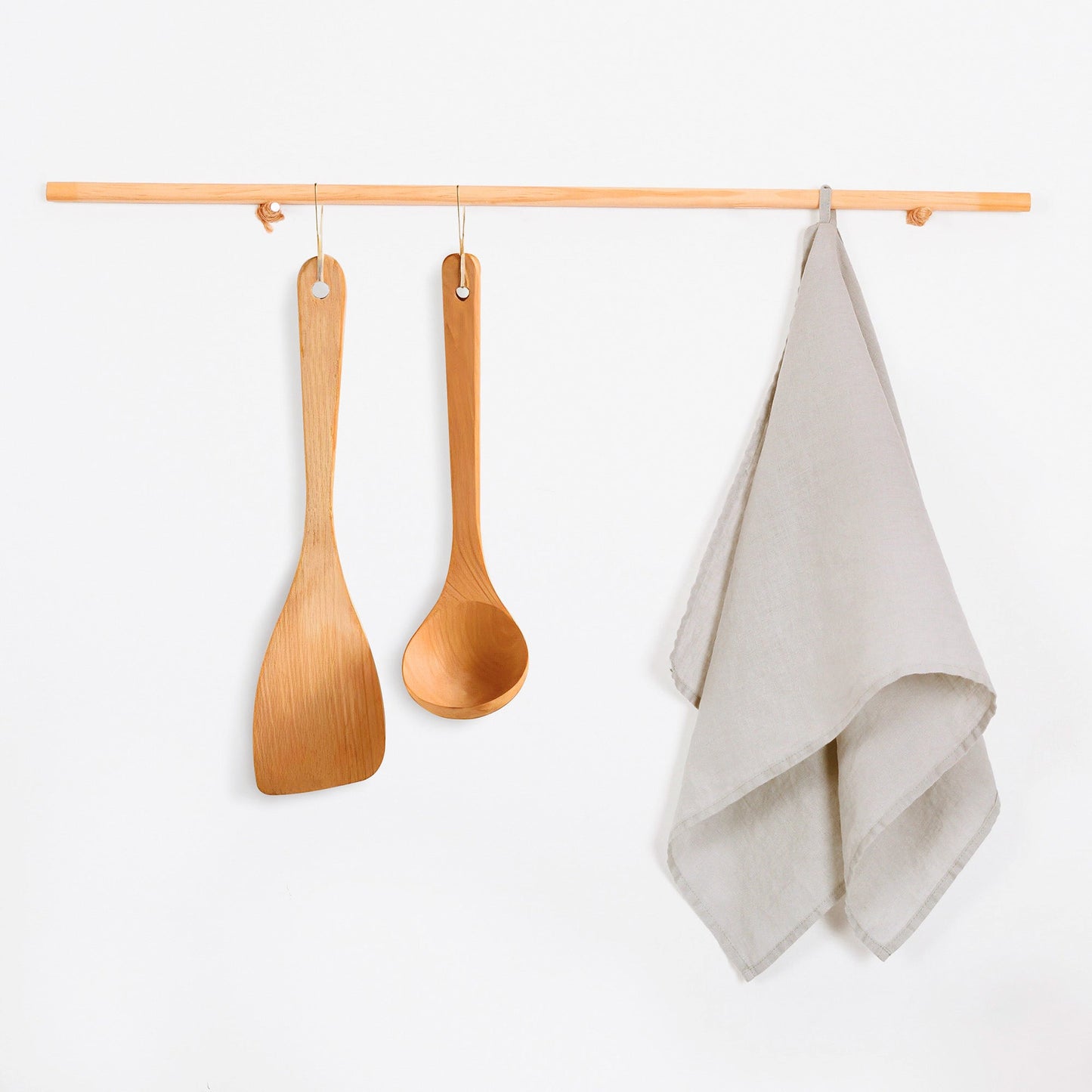 A light natural wood hanging rack with two wooden utensils and a folded beige tea towel, shown against a plain white wall.