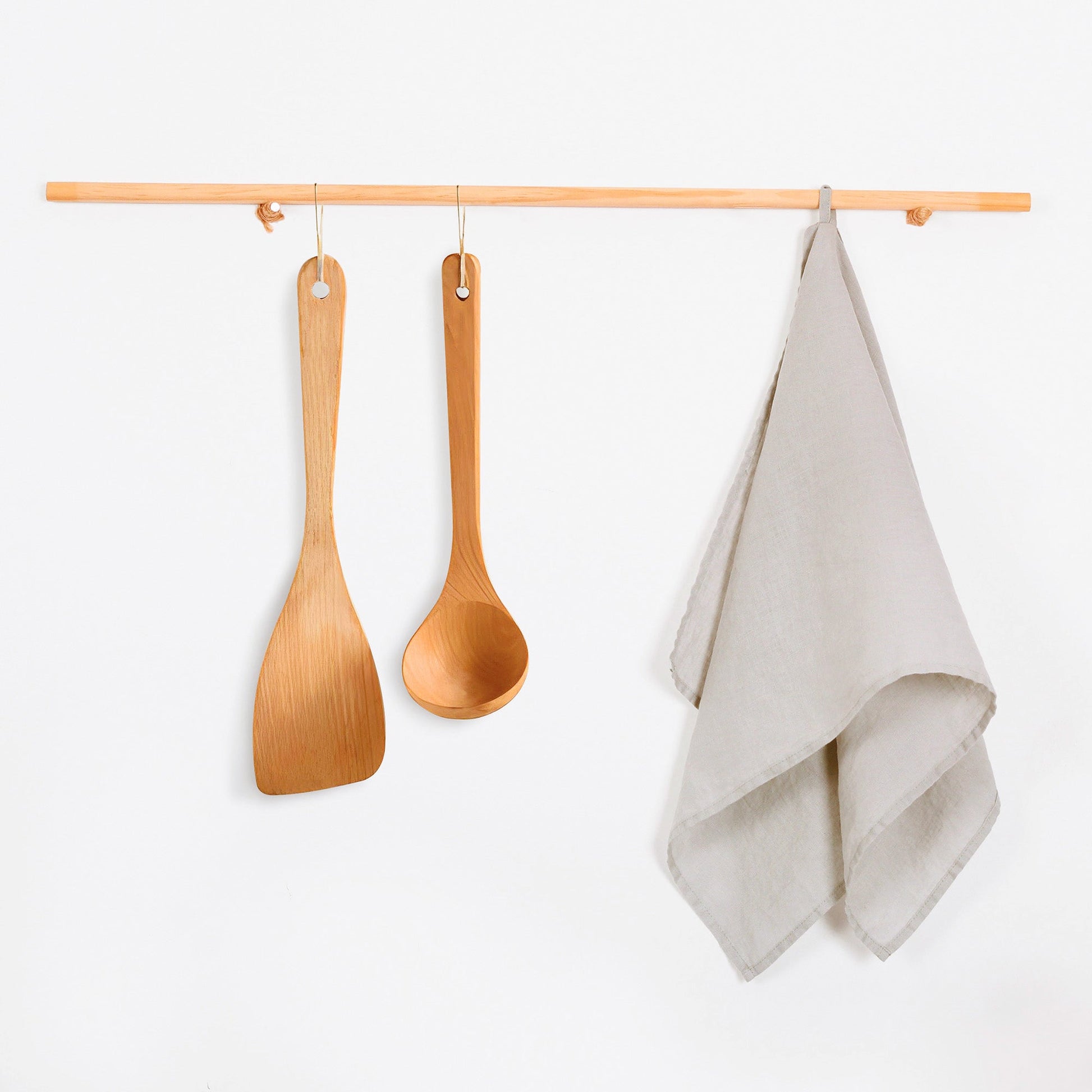 A light natural wood hanging rack with two wooden utensils and a folded beige tea towel, shown against a plain white wall.