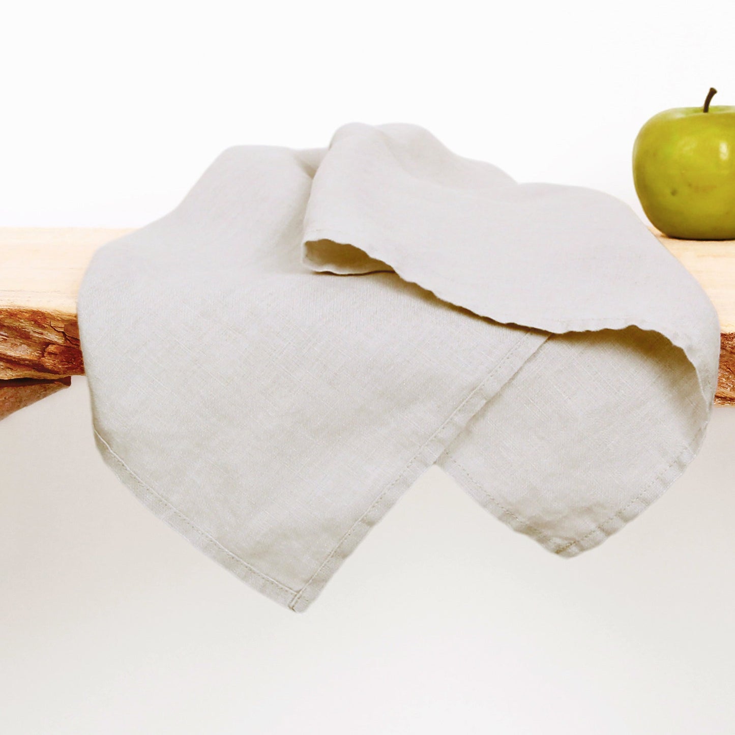 A light beige linen tea towel draped over a rustic wooden slice, with a green apple visible in the background on a white surface.