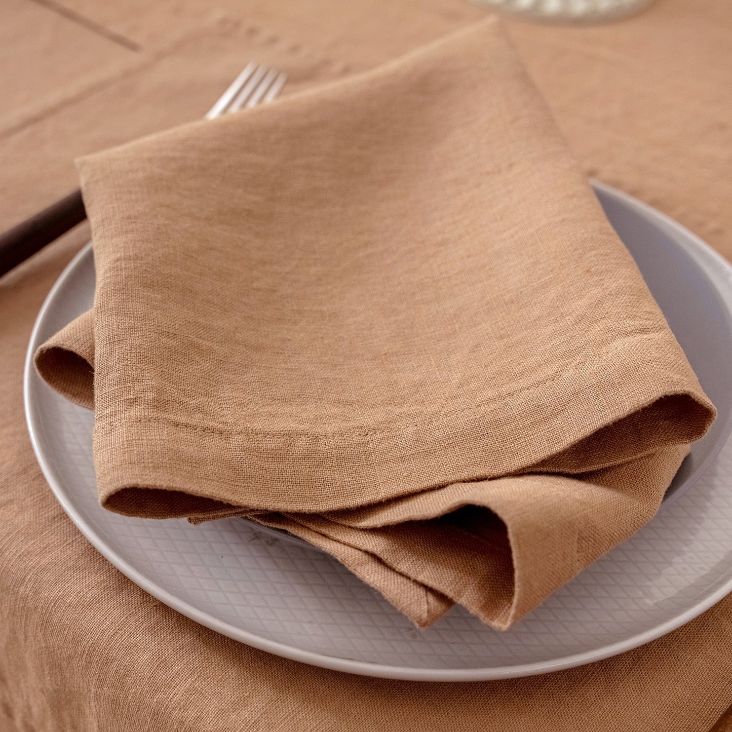 A folded tan linen napkin with a subtle woven texture, neatly arranged on a white plate with a fork, set against a matching tan linen tablecloth.