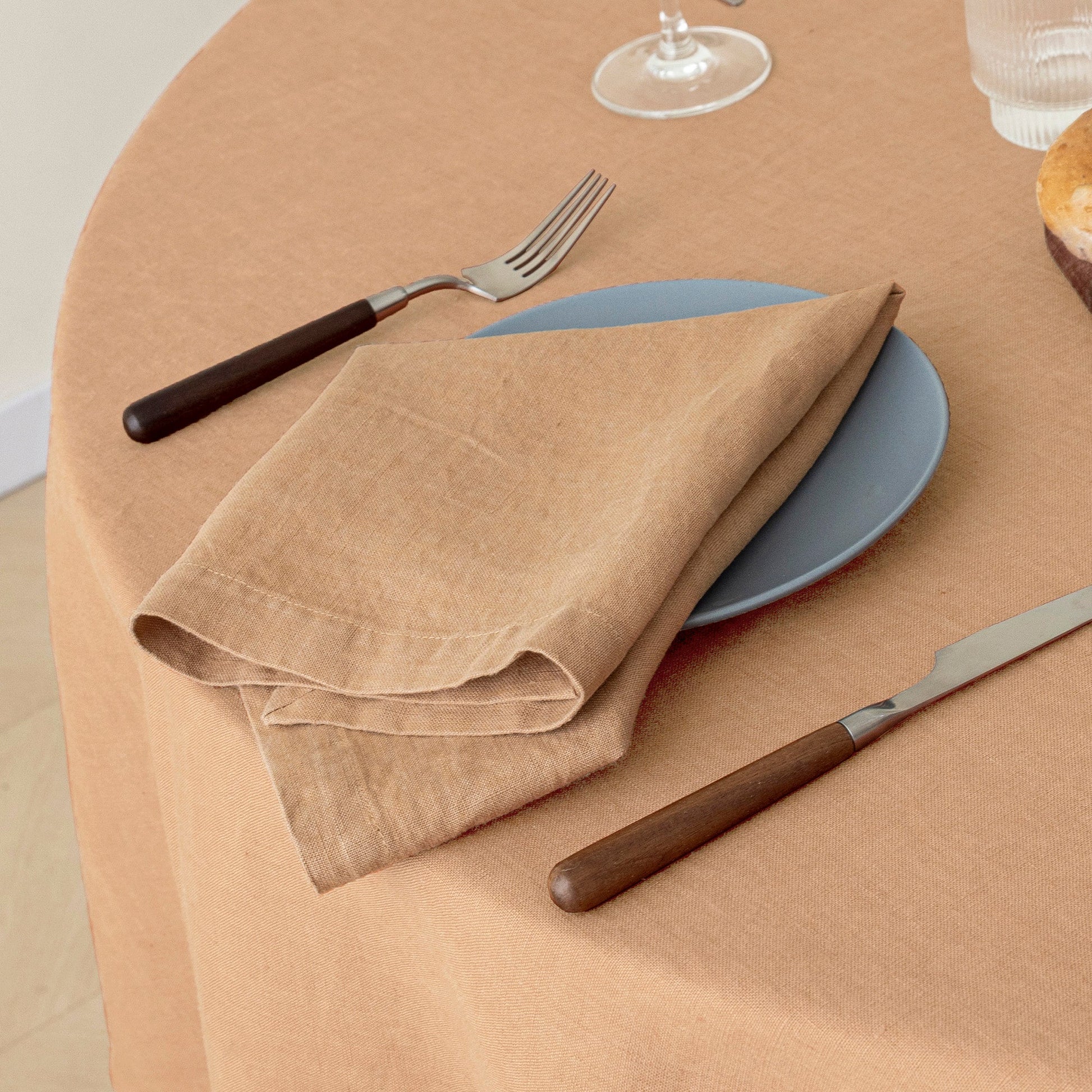 A beige linen napkin folded on a blue plate with a fork and knife featuring dark brown handles, set on a peach-colored tablecloth.