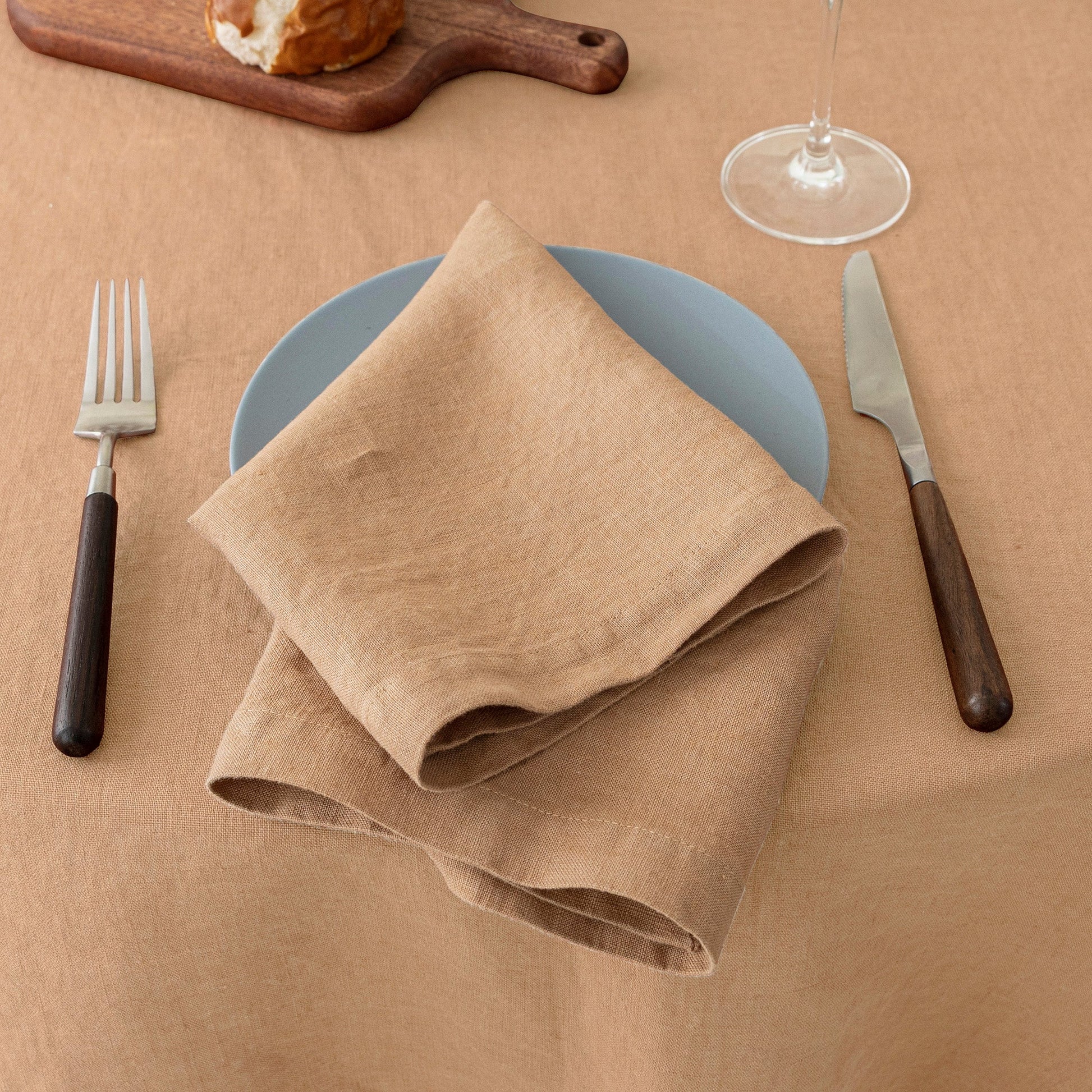 A folded beige linen napkin on a light blue plate, styled on a beige tablecloth with a blue plate, silver and dark wood cutlery, a wine glass base, and a wooden cutting board in the background.