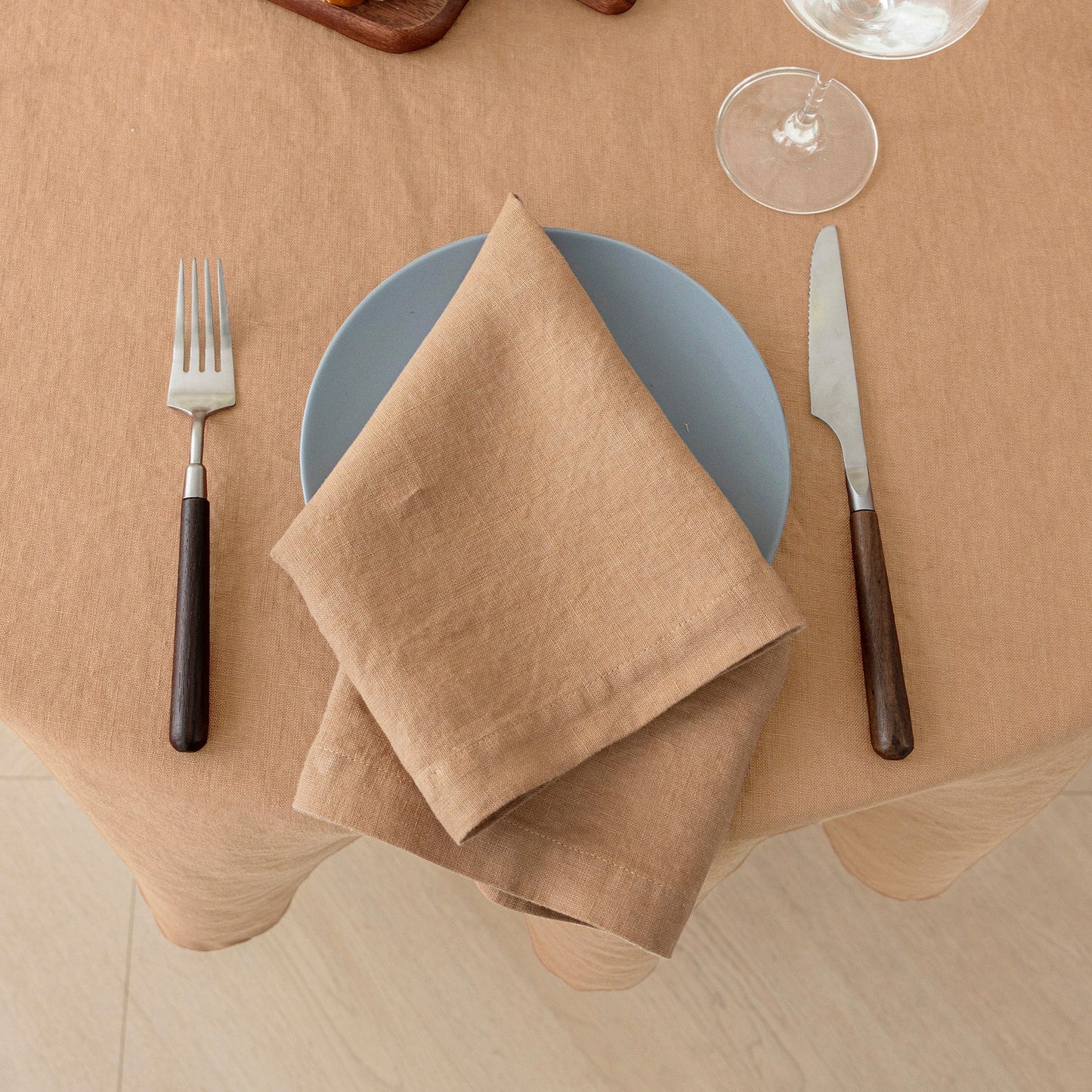 A beige linen napkin folded over a light blue plate on a beige tablecloth, styled with a fork and knife on a dining table.