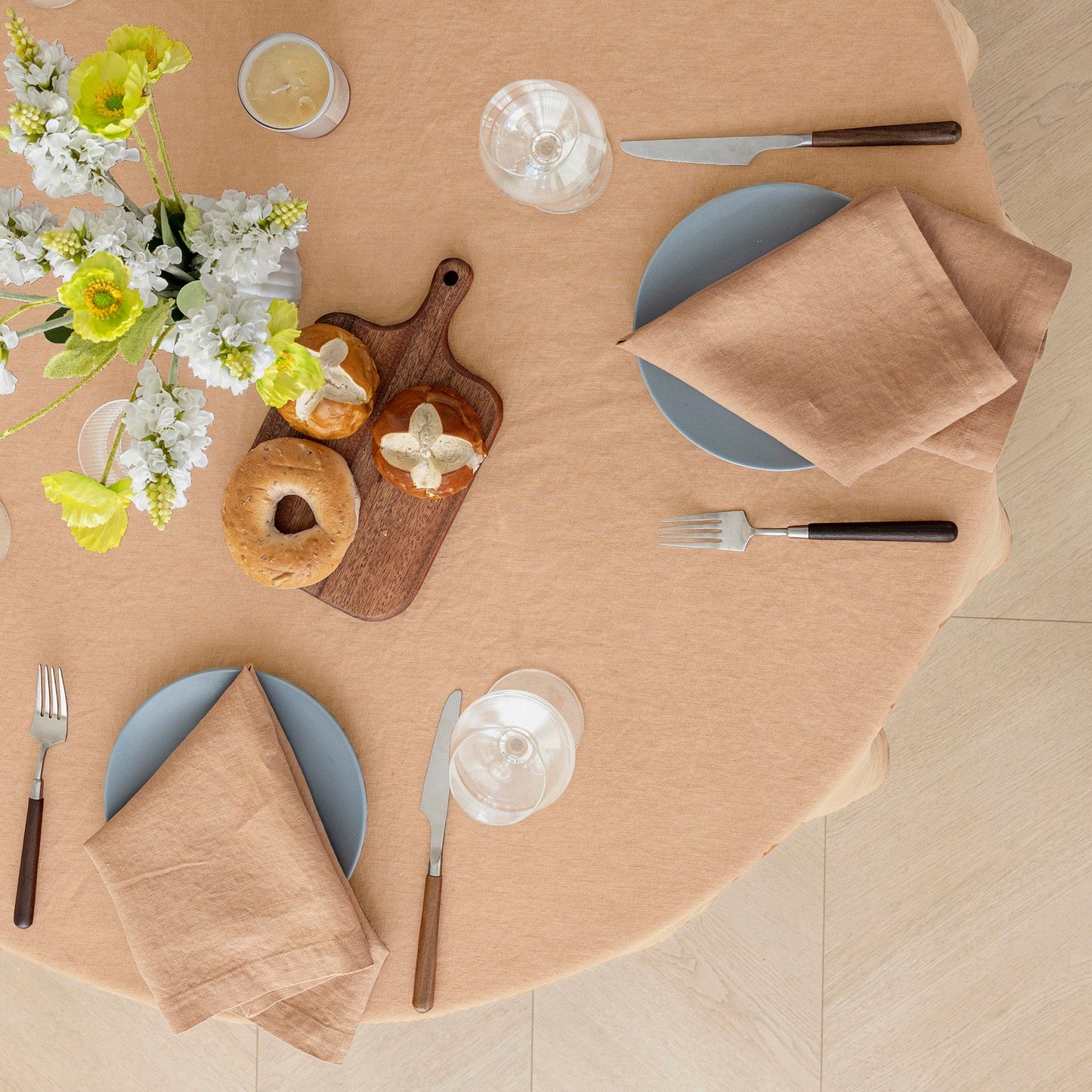 An overhead flat-lay of a round table set with a blush pink tablecloth, blue plates, beige napkins, silver cutlery, a wooden cutting board with pastries, and a floral centerpiece on a light wood floor.