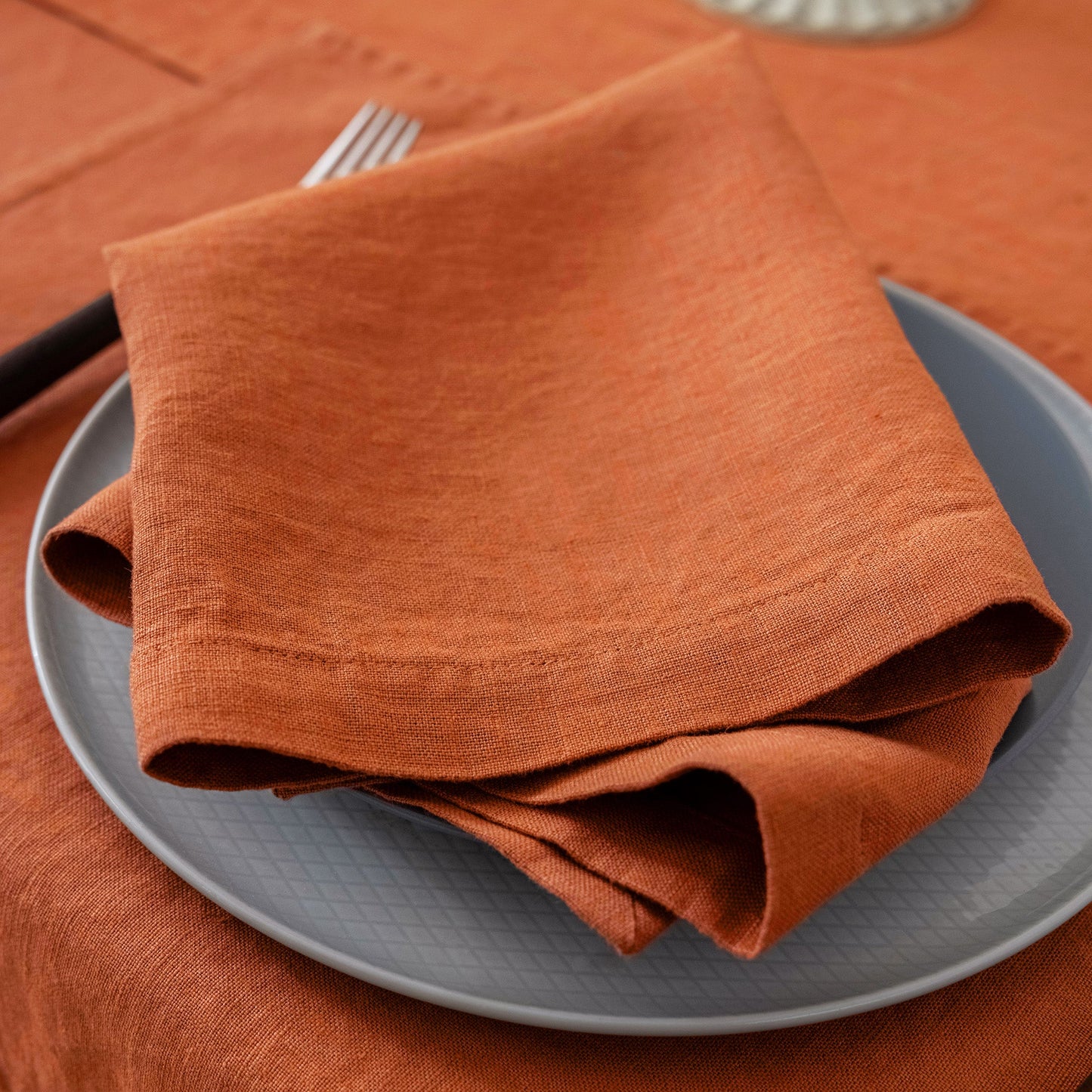An orange-brown linen napkin folded on a gray plate, set on an orange tablecloth with a fork partially visible.