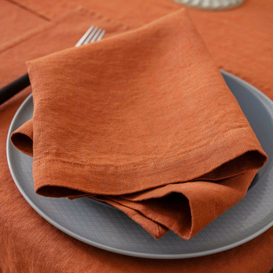 An orange-brown linen napkin folded on a gray plate, set on an orange tablecloth with a fork partially visible.