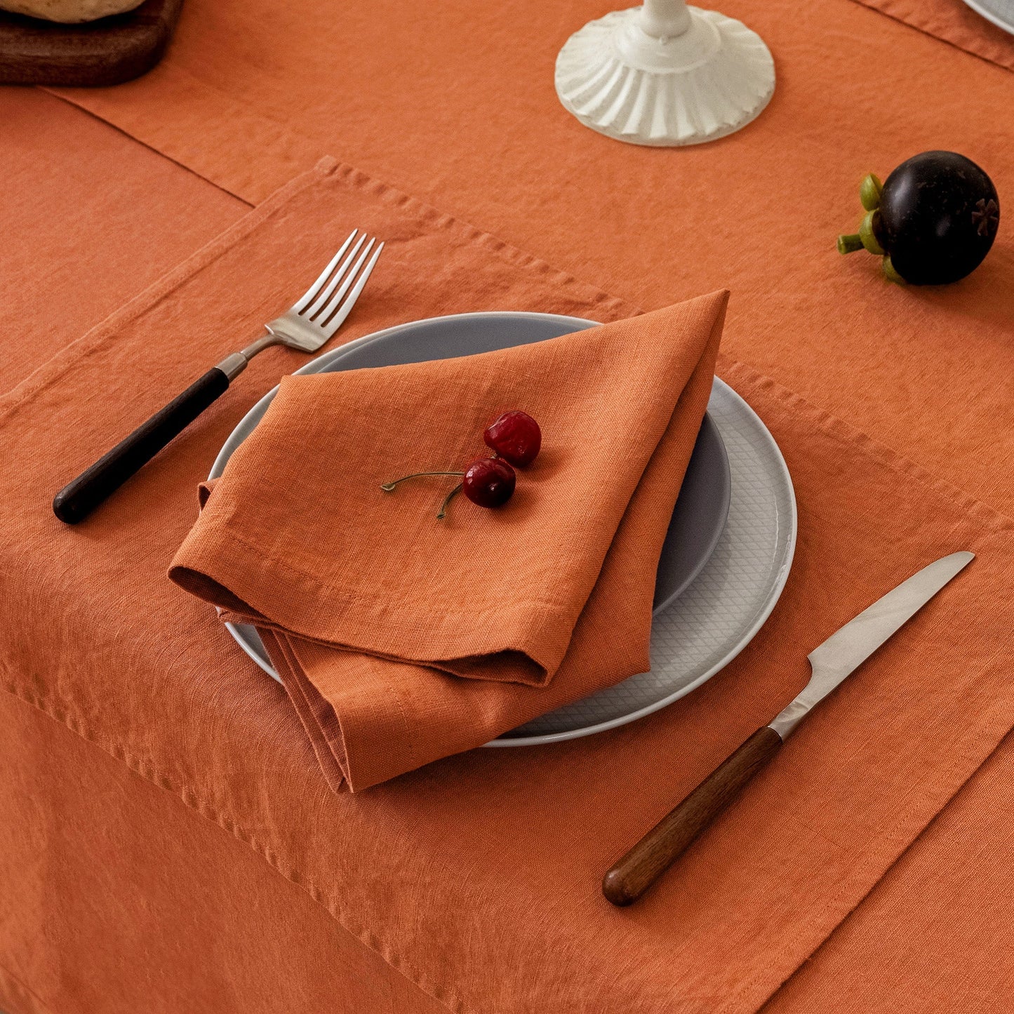 An orange linen napkin folded on top of a gray plate, set on an orange tablecloth with a fork, knife, and a small dark fruit nearby.