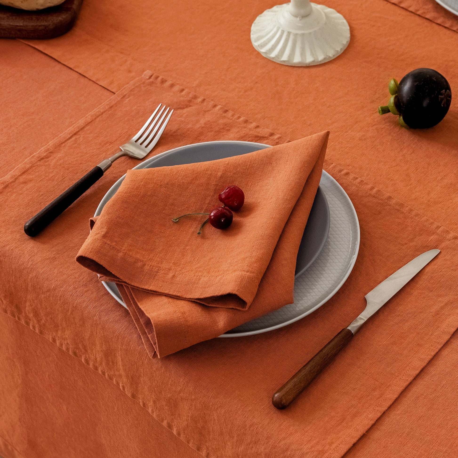 An orange linen napkin folded on top of a gray plate, set on an orange tablecloth with a fork, knife, and a small dark fruit nearby.