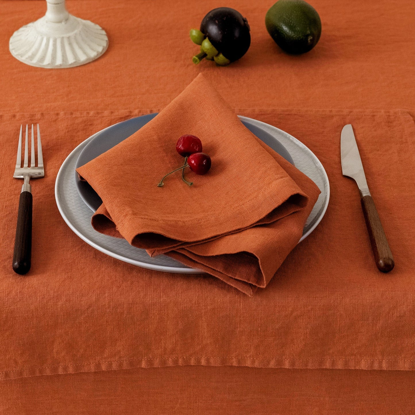 An orange linen tablecloth with matching placemats, set with a gray plate, orange napkin, cherry garnish, silver fork, and knife, with fruit and a white candle base in the background.