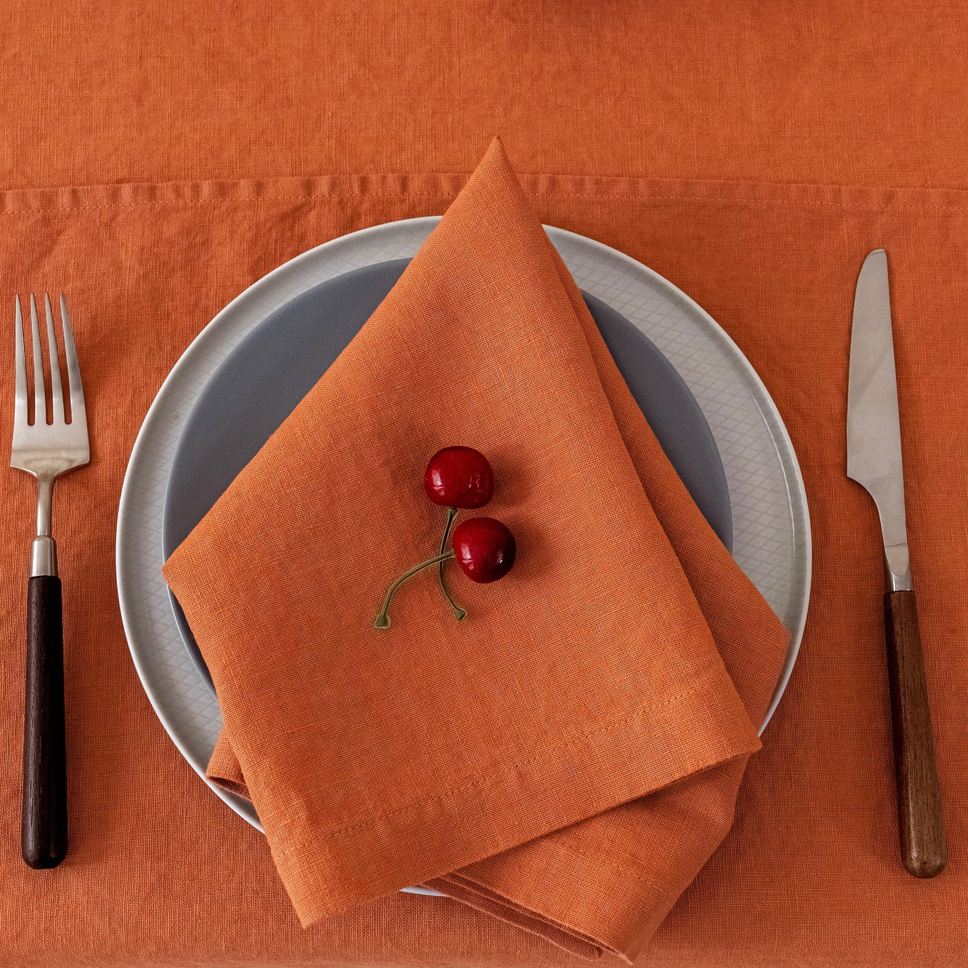 An orange linen napkin folded into a triangle and placed on top of a gray plate, set on an orange tablecloth with a fork on the left and a knife on the right. Two small red cherries rest on the napkin.