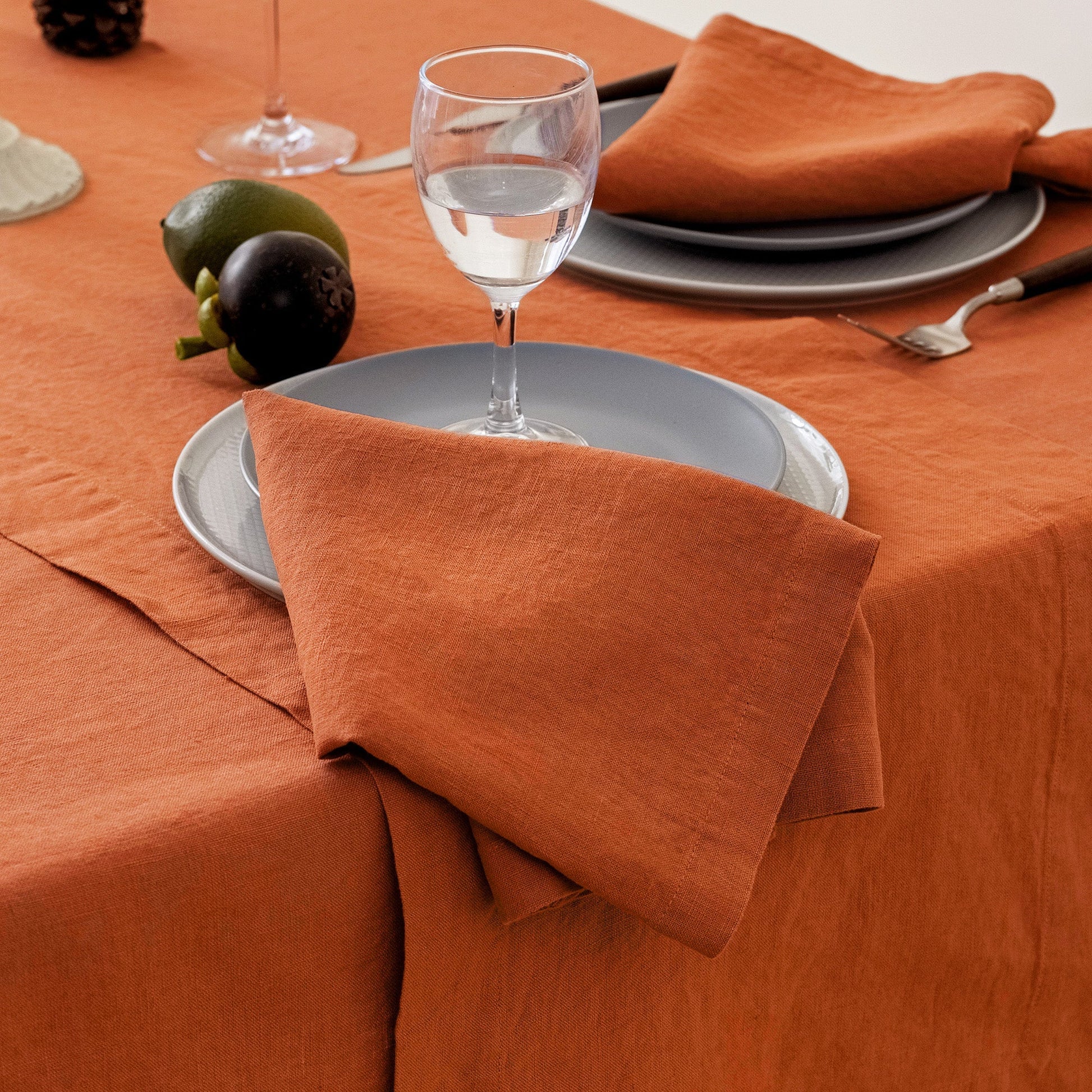 An orange linen tablecloth and matching folded napkins on a styled dining table with gray plates, a wine glass, and small green and purple fruits.
