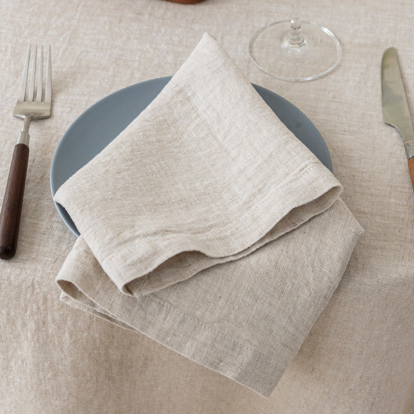Folded beige napkin on a gray plate with cutlery on a beige tablecloth.