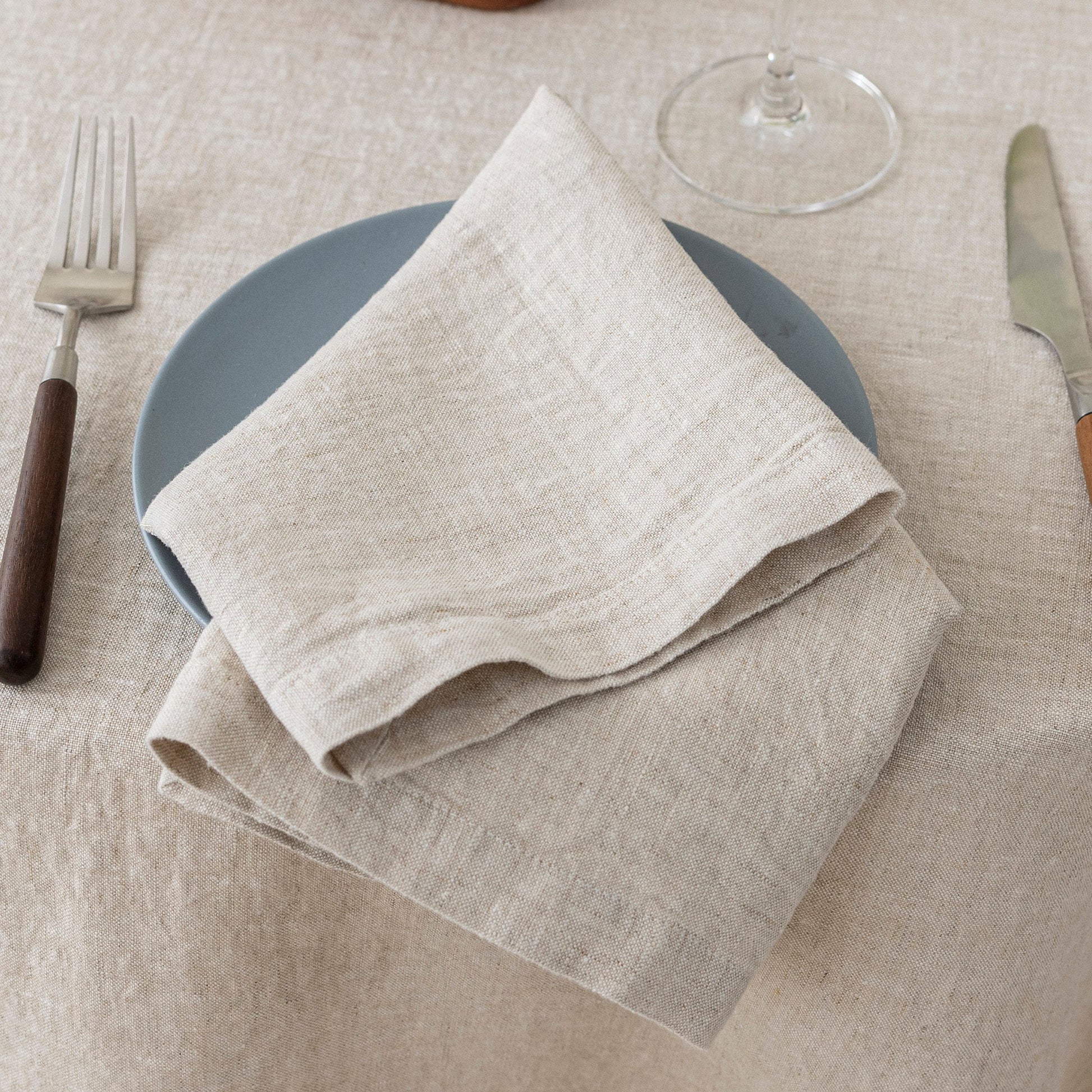 Folded beige napkin on a gray plate with cutlery on a beige tablecloth.