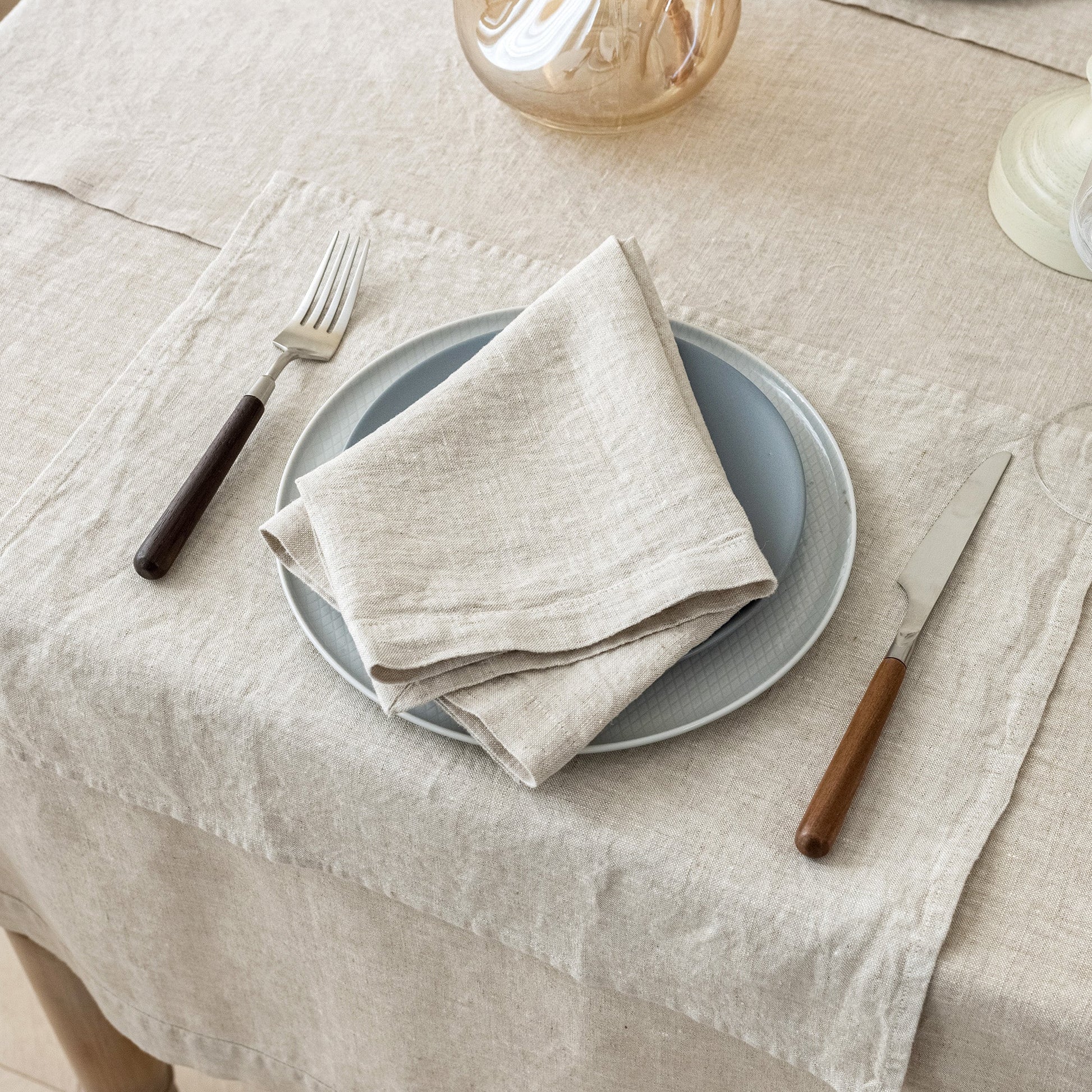 A styled tabletop setting with a light beige linen tablecloth, a folded beige napkin on a blue-gray plate, a fork with a dark handle, and a knife with a brown handle.