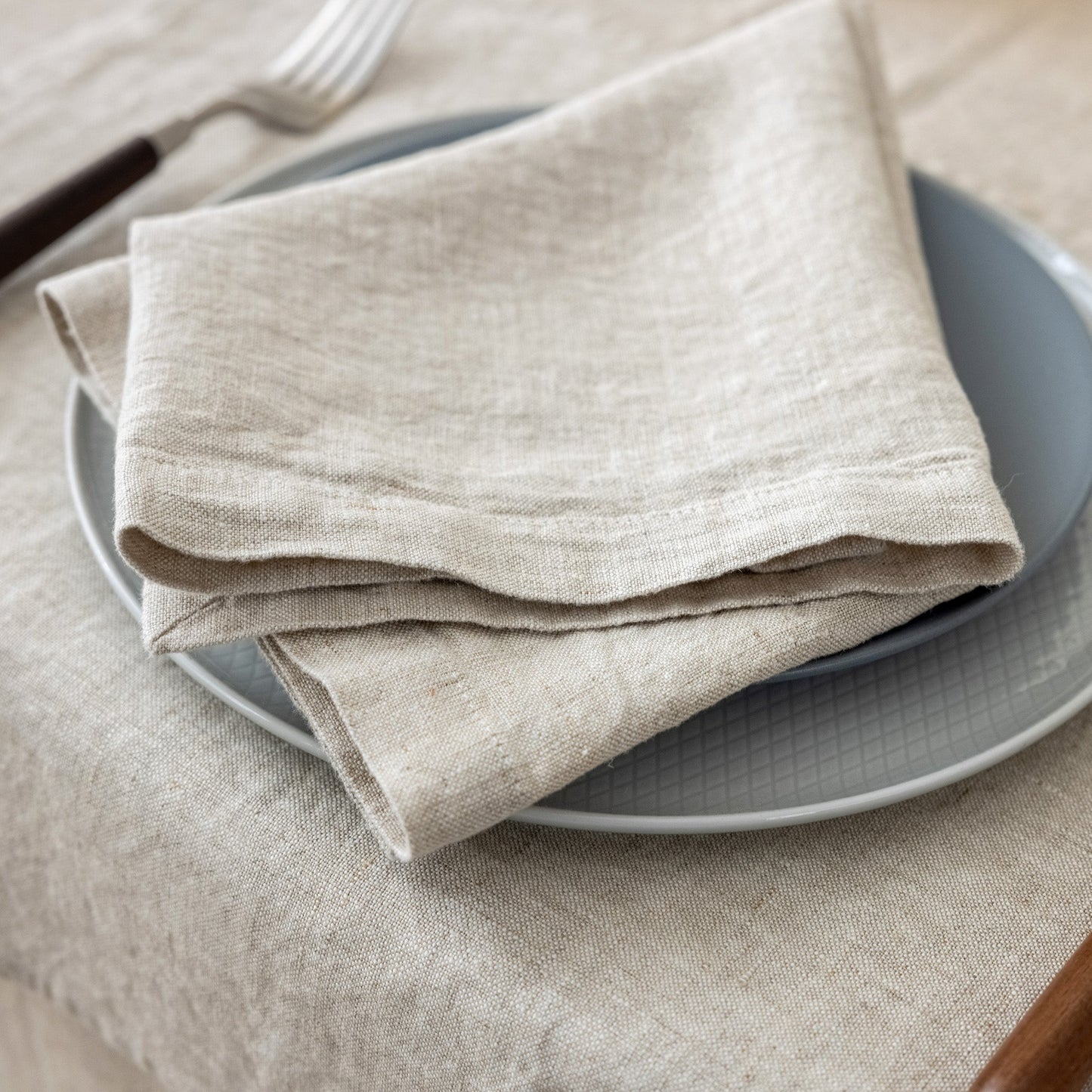 A folded beige linen napkin with a natural slub texture, layered over a gray plate on a matching beige tablecloth, with a fork partially visible in the background.