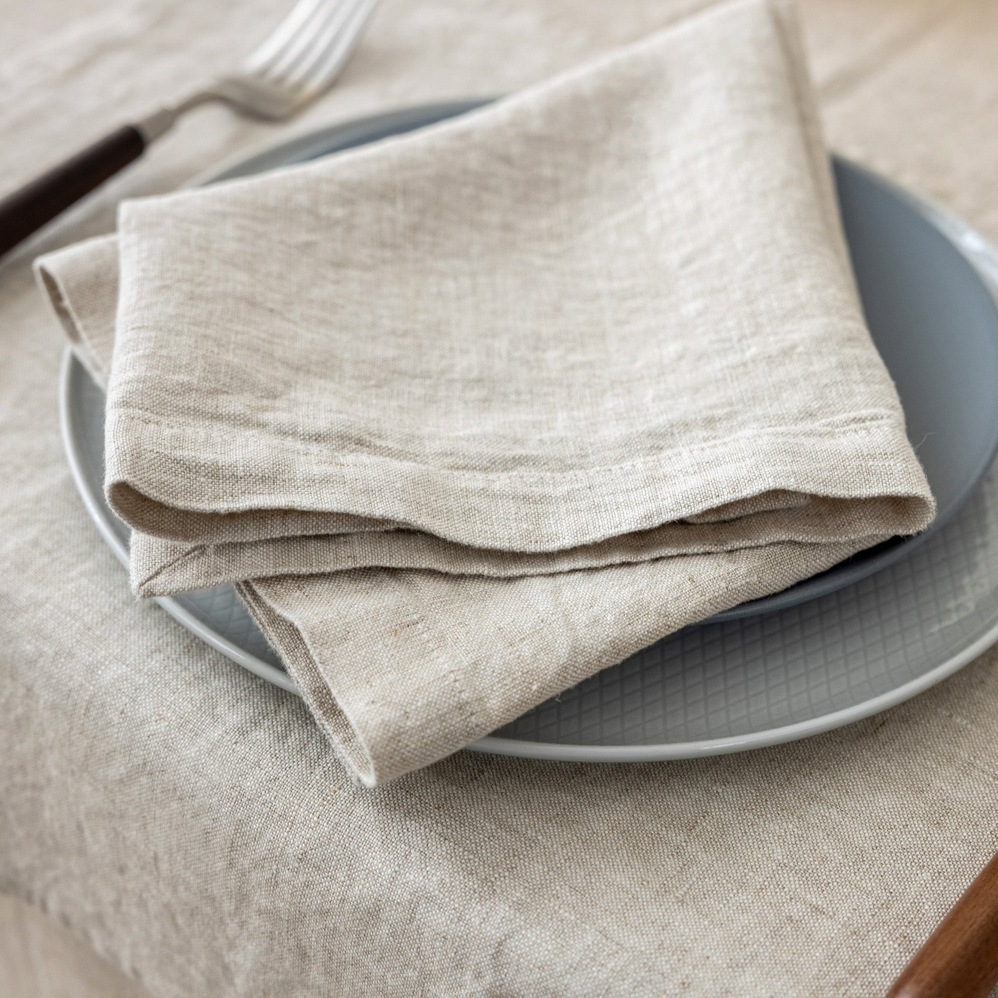 A folded beige linen napkin with a natural slub texture, layered over a gray plate on a matching beige tablecloth, with a fork partially visible in the background.