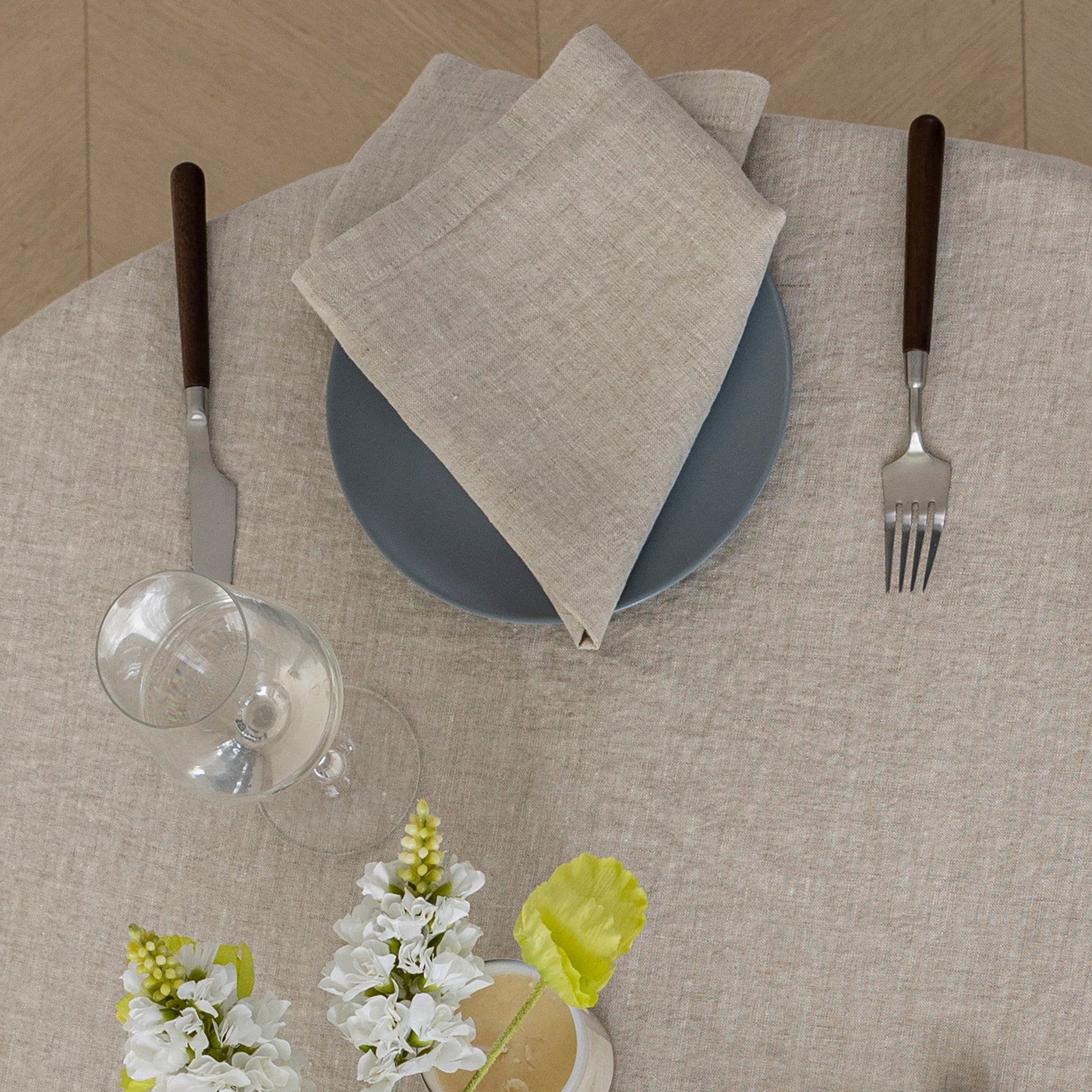 A styled table setting featuring a beige linen tablecloth, a folded beige napkin on a slate blue plate, a silver knife and fork with dark brown handles, a wine glass, and small white flowers.