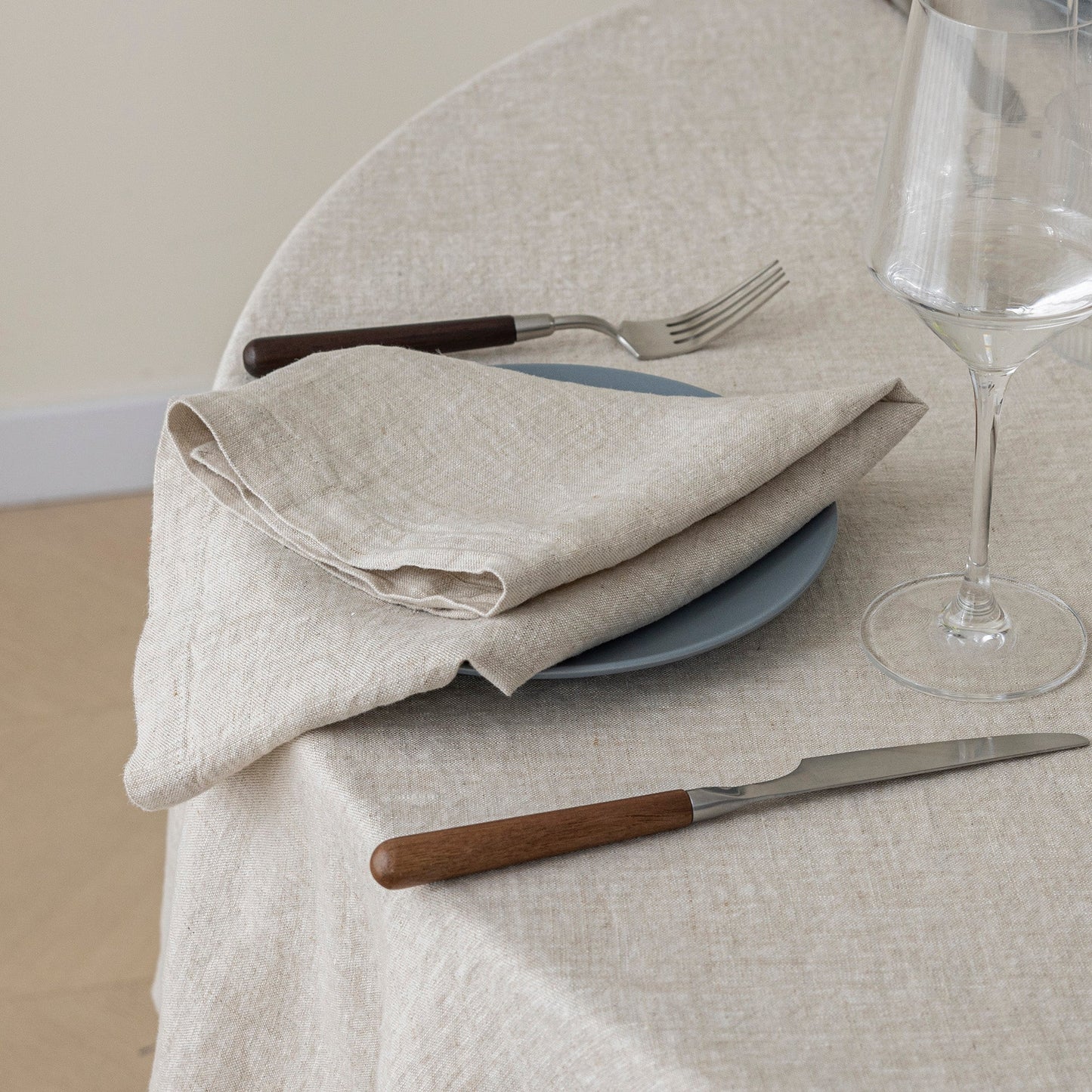 A folded beige linen napkin on a blue plate with a fork and knife on a round table covered in a matching beige tablecloth. A wine glass is partially visible on the right.