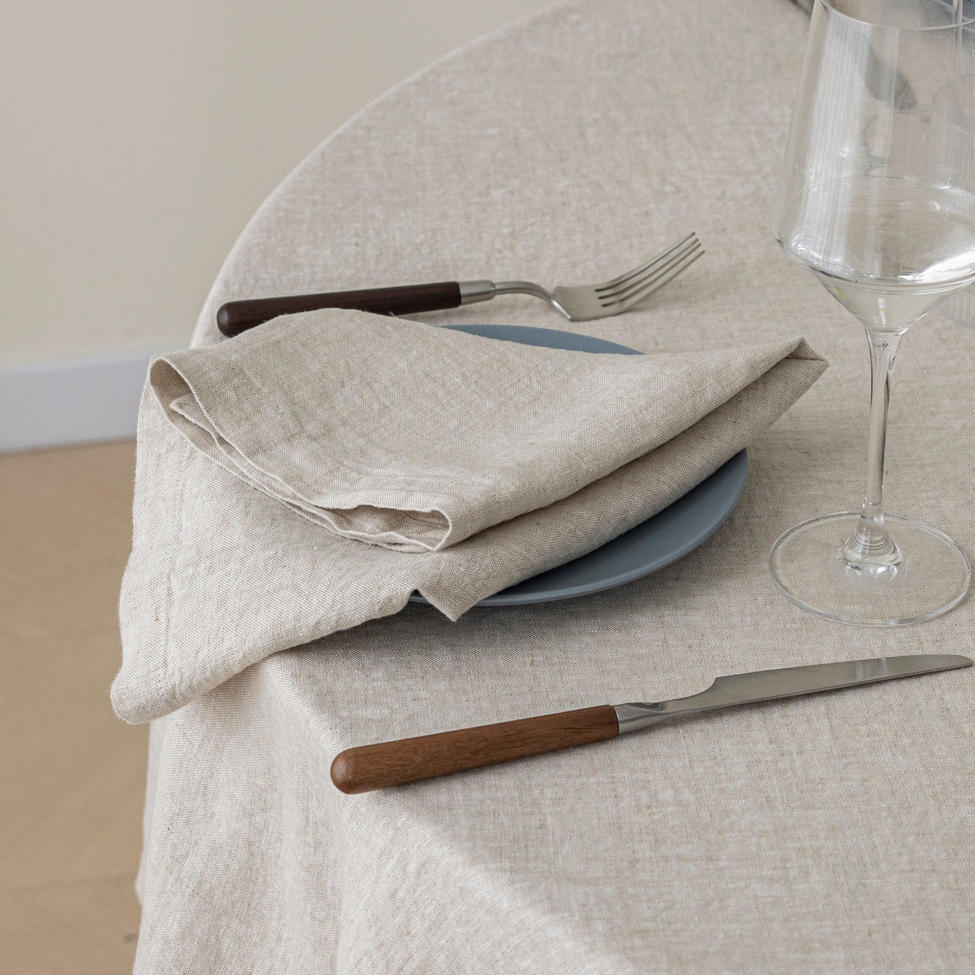 A folded beige linen napkin on a blue plate with a fork and knife on a round table covered in a matching beige tablecloth. A wine glass is partially visible on the right.