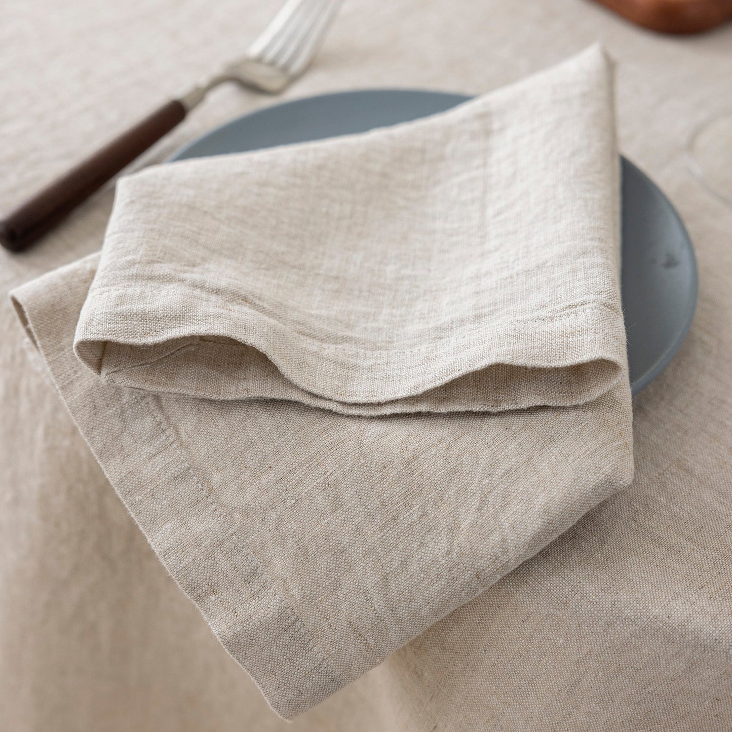 A folded beige linen napkin with a subtle woven texture, styled on a dark gray plate over a matching beige tablecloth with a fork in the background.