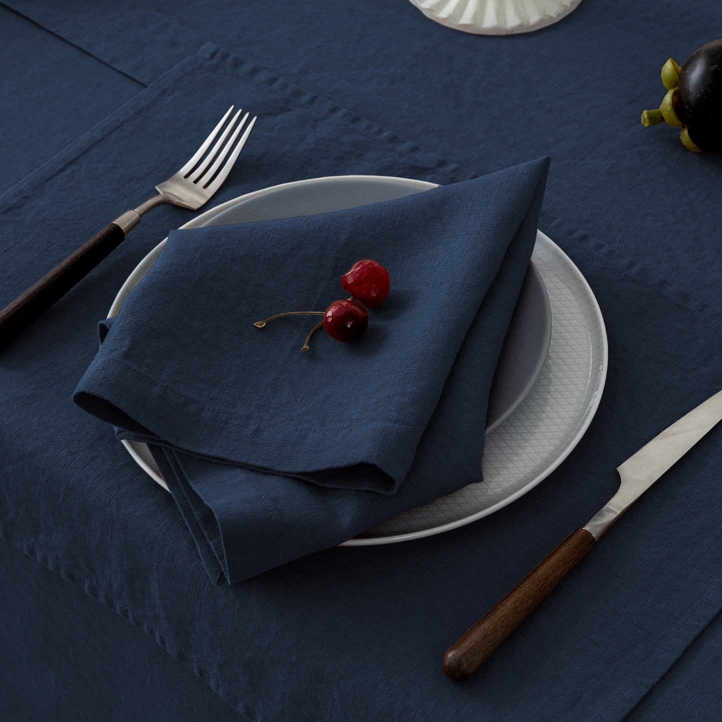Napkin folded on a plate with cherries, fork, knife, and glass on a dark blue tablecloth.