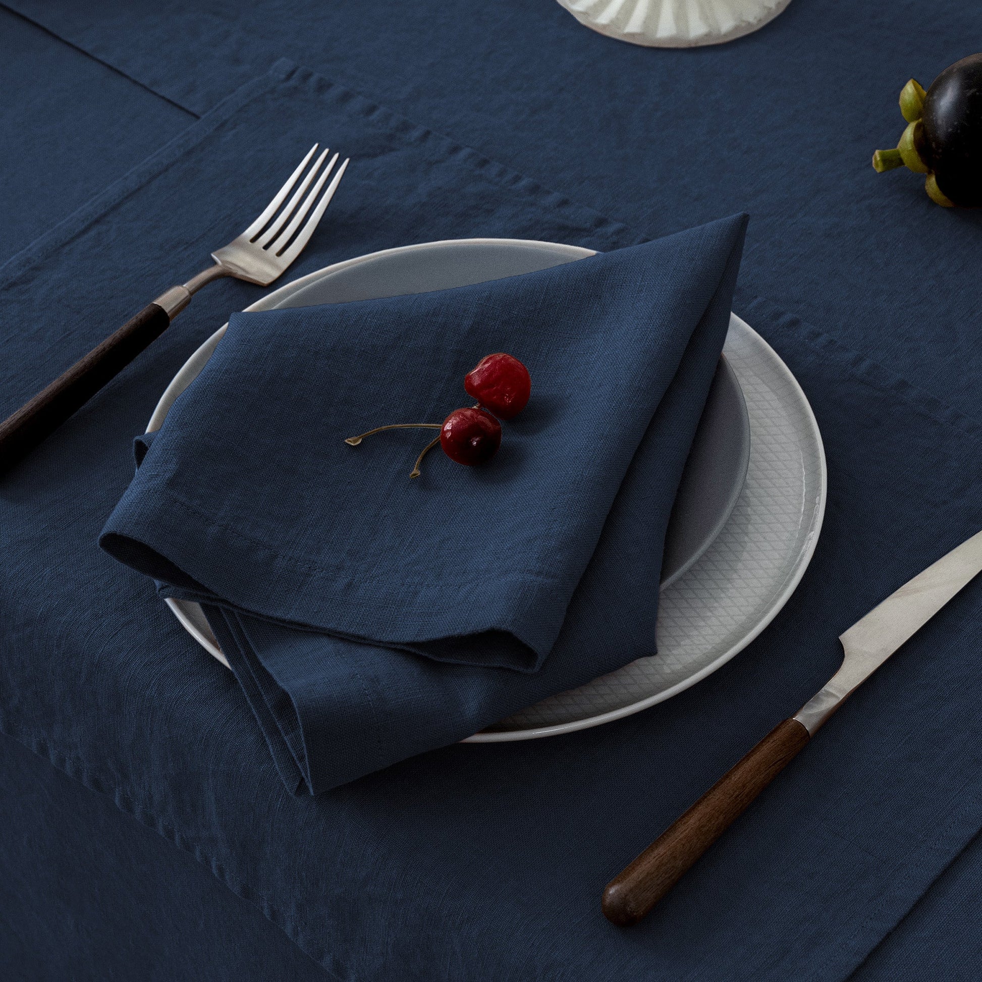 Napkin folded on a plate with cherries, fork, knife, and glass on a dark blue tablecloth.