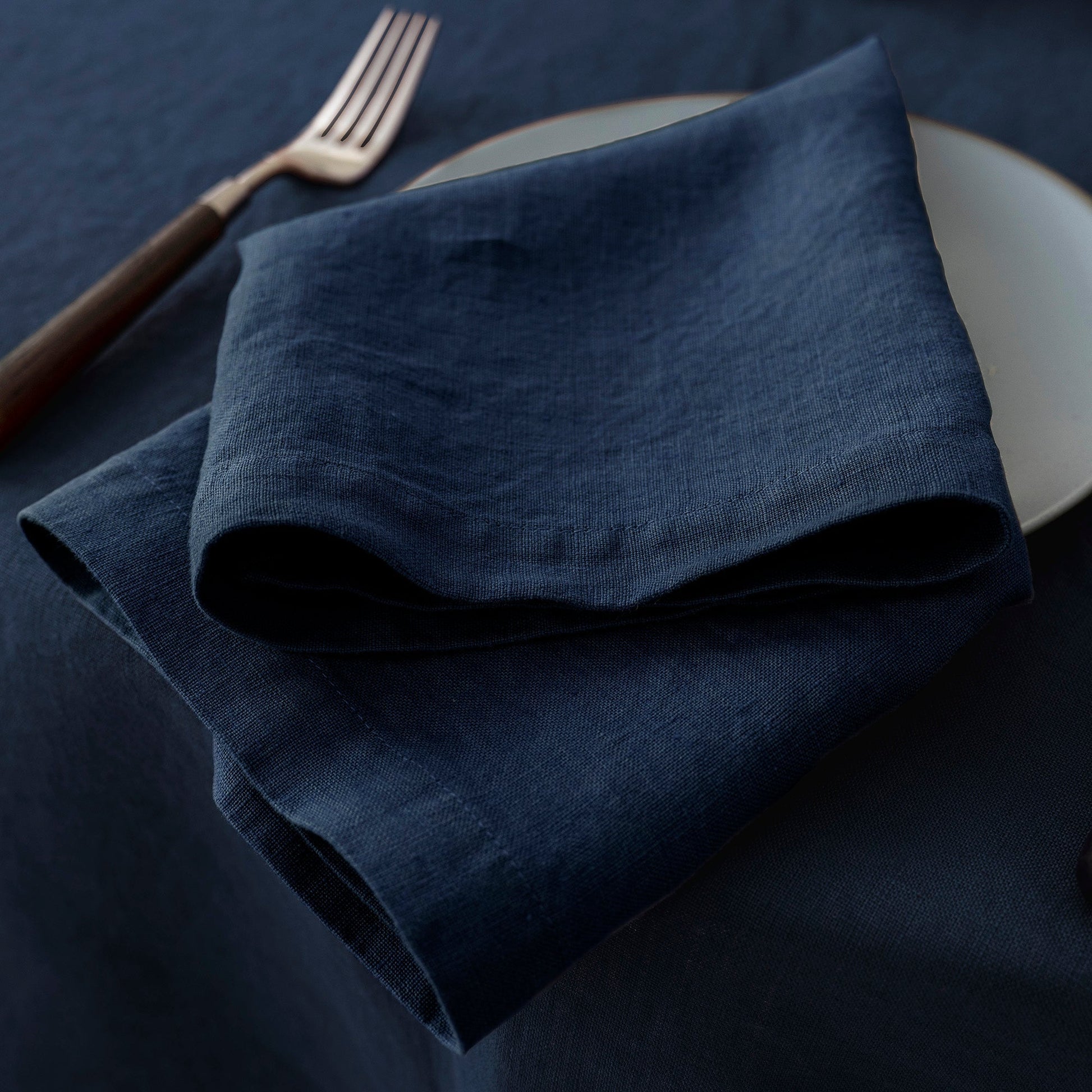 A folded dark navy blue linen napkin on a matching dark navy tablecloth, styled with a white plate and a fork with a dark wooden handle.