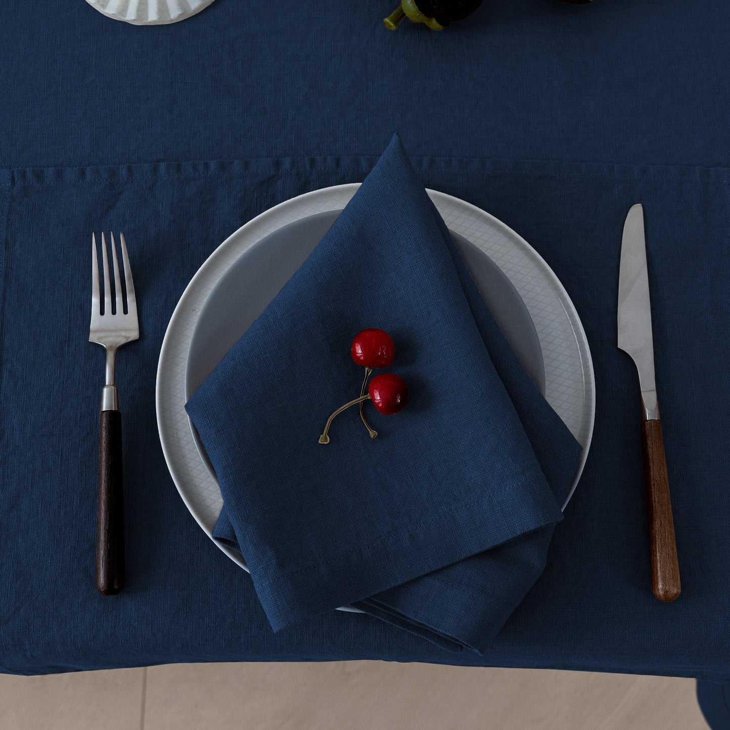 A styled table setting featuring a navy blue tablecloth, matching navy napkin, white plates, silver fork and knife with dark and wood handles, and two red cherries on the napkin.