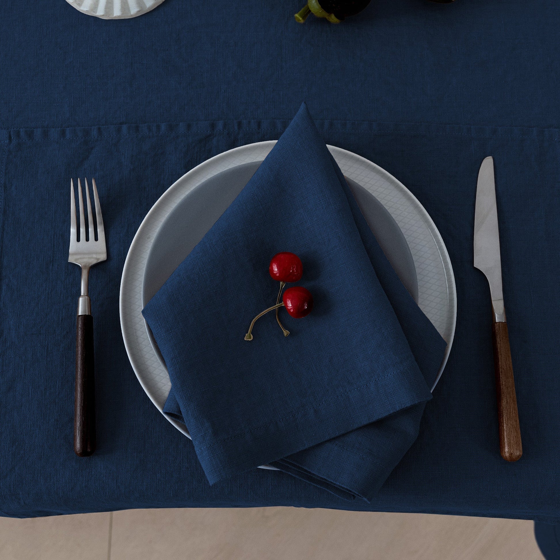 A styled table setting featuring a navy blue tablecloth, matching navy napkin, white plates, silver fork and knife with dark and wood handles, and two red cherries on the napkin.