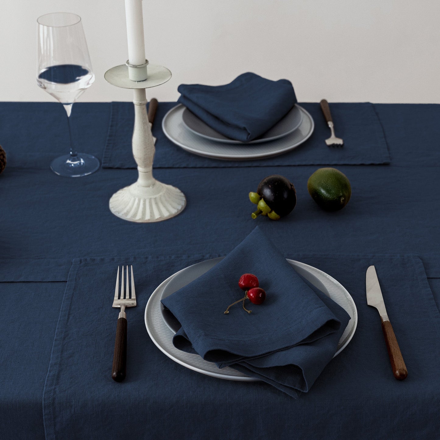A styled dining table setting featuring a dark navy blue tablecloth, matching napkins, white plates, silver cutlery with dark handles, a wine glass, and a white candlestick, accented with small red cherries and green fruit.