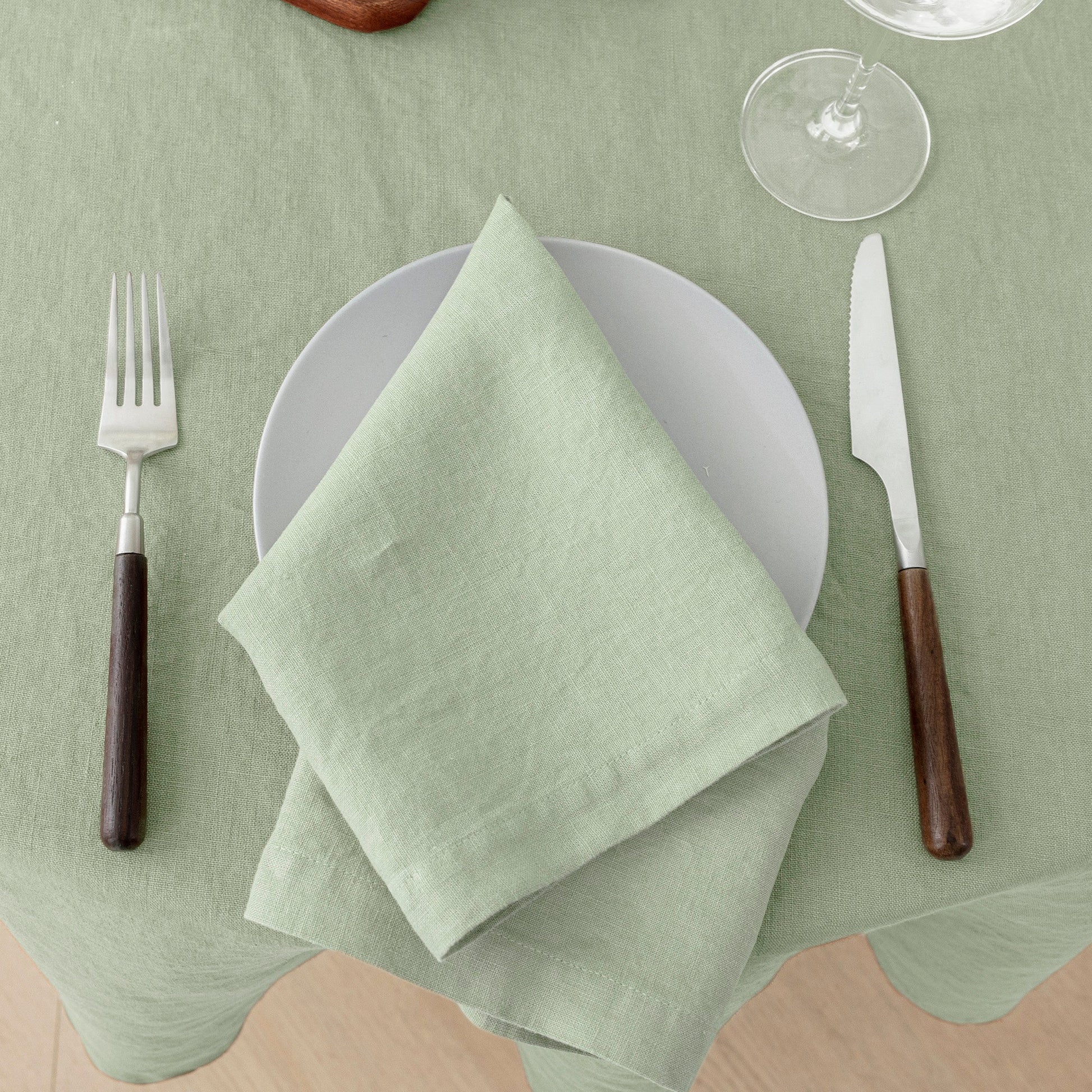 Green table setting with a folded napkin on a white plate, fork, knife, and wine glass on a green tablecloth.