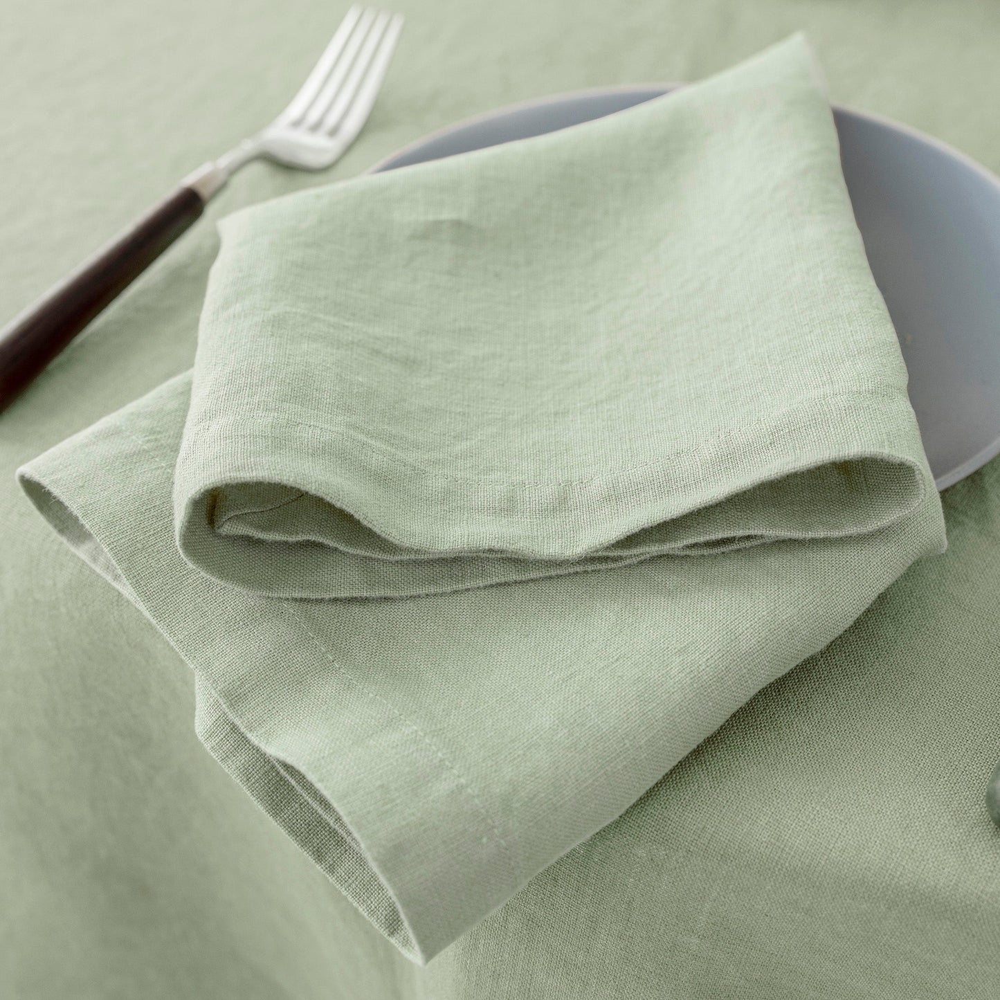 A folded light sage green linen napkin on a matching tablecloth, styled with a gray plate and a fork with a dark brown handle.