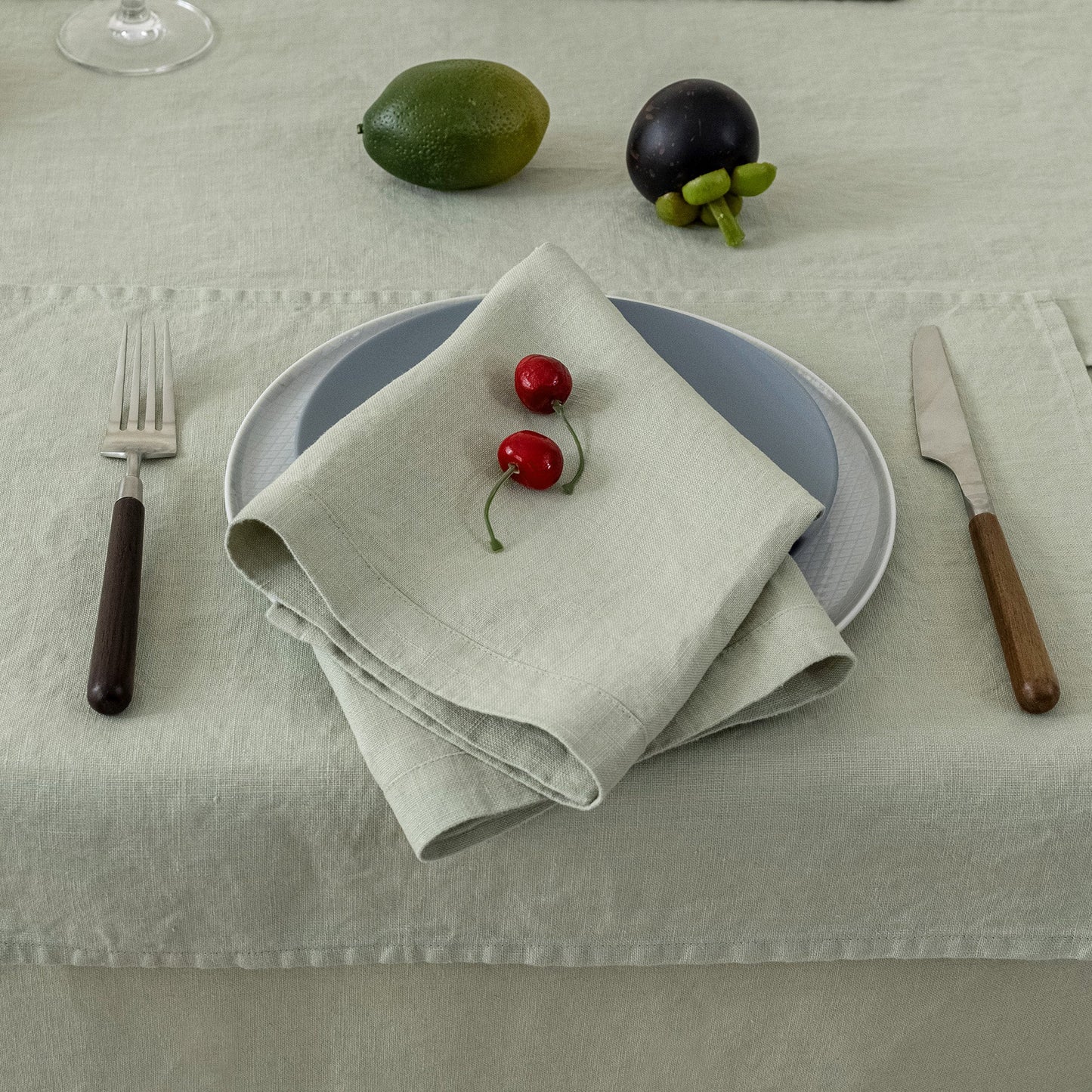 A styled table setting featuring a folded light beige linen napkin with two red cherries on top, layered over a blue-gray plate and a white plate, set on a textured light beige tablecloth. A silver fork with a dark handle, a knife with a wooden handle, a green lime, and a dark purple fruit are arranged around it.
