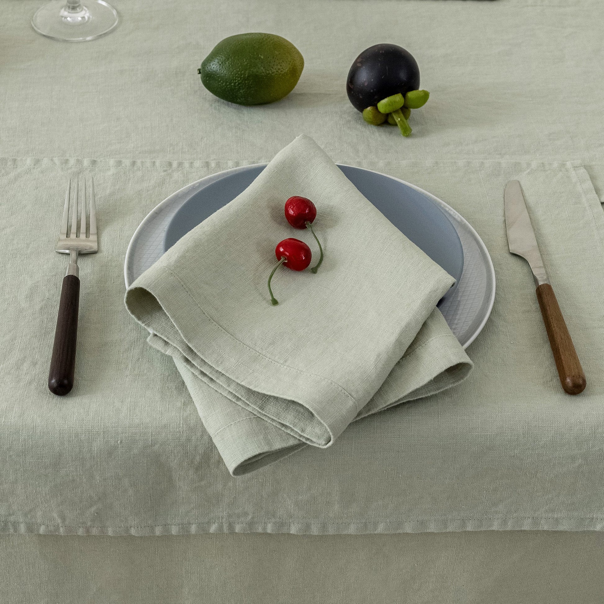 A styled table setting featuring a folded light beige linen napkin with two red cherries on top, layered over a blue-gray plate and a white plate, set on a textured light beige tablecloth. A silver fork with a dark handle, a knife with a wooden handle, a green lime, and a dark purple fruit are arranged around it.