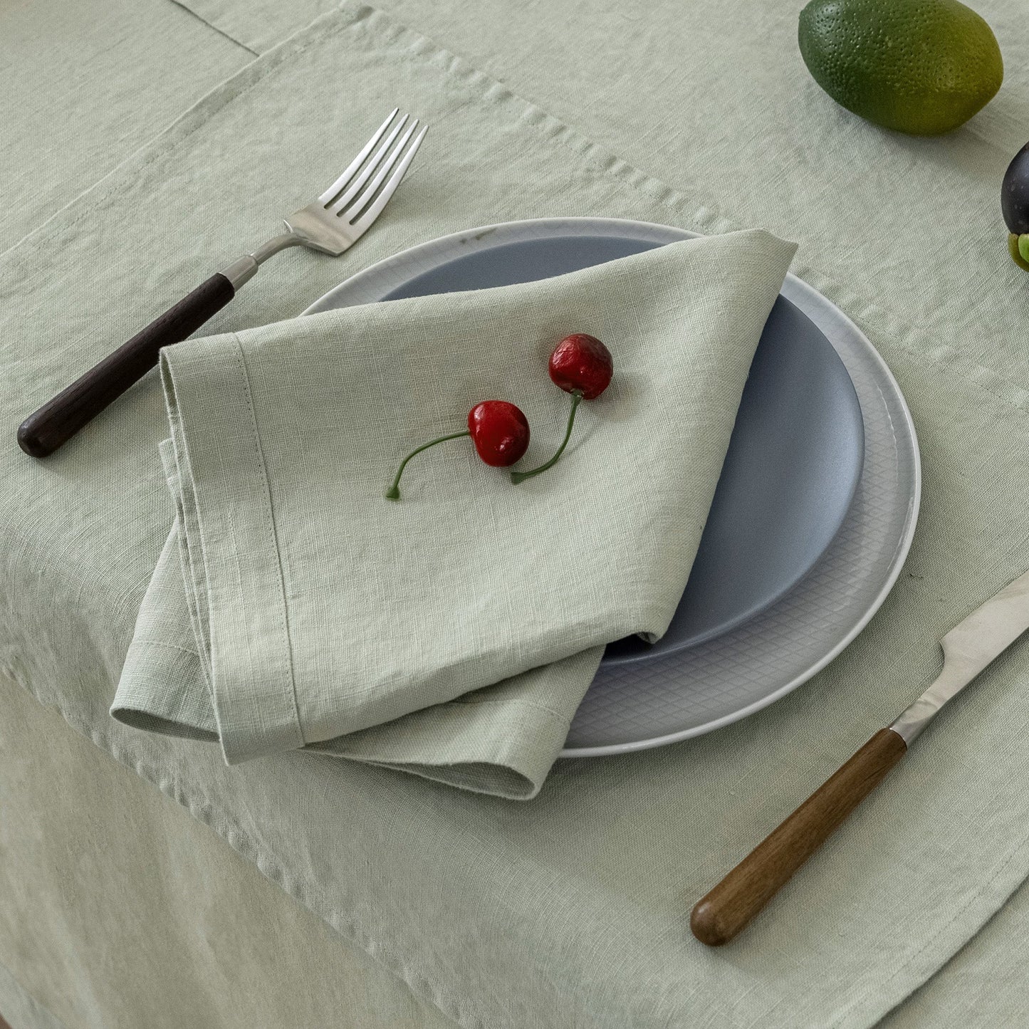 A folded light beige linen napkin with two red cherries on it, layered over a light blue-gray plate, set on a matching tablecloth with a fork and knife nearby.