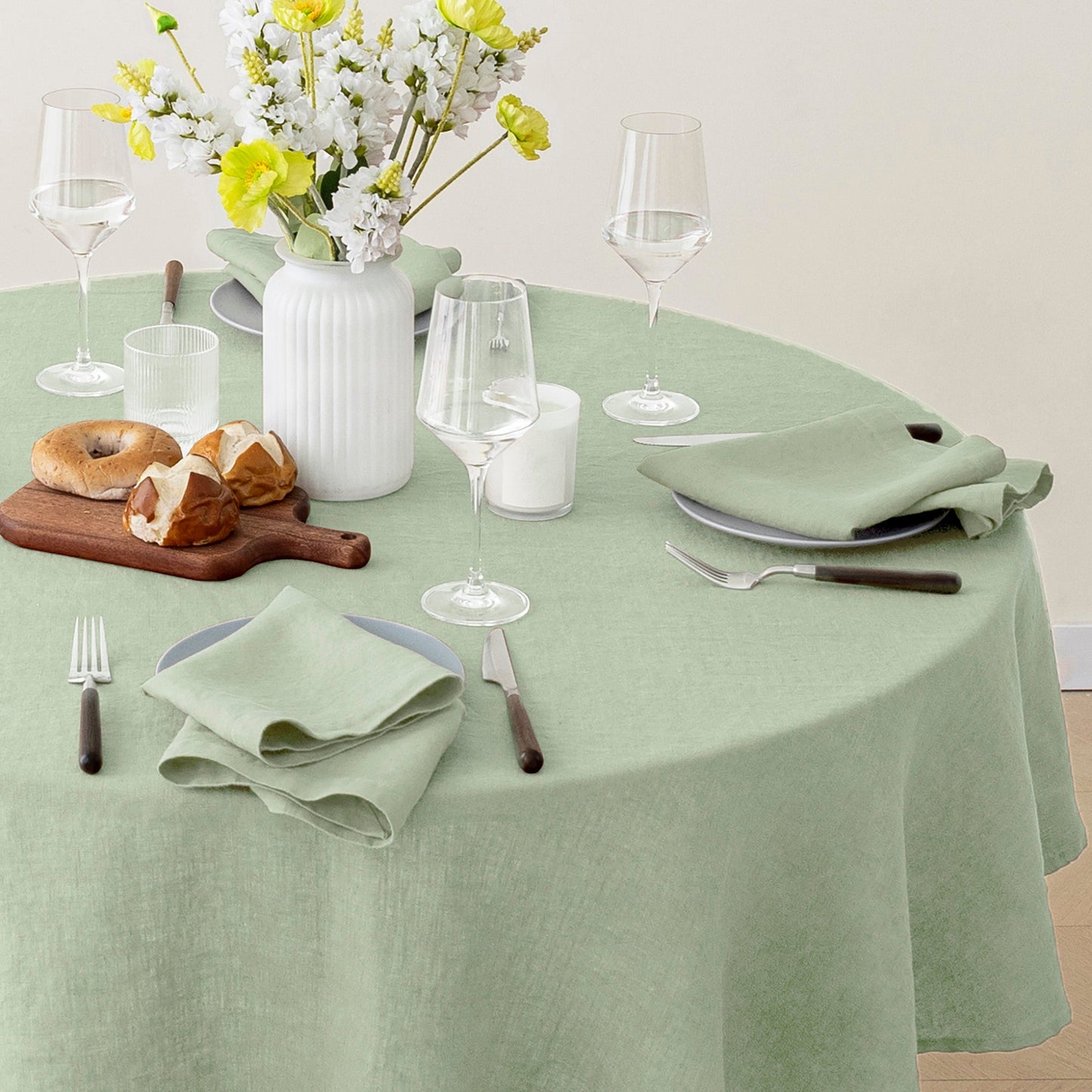 A sage green linen tablecloth draped over a round dining table, styled with white plates, folded napkins, wine glasses, a wooden cutting board with bagels, and a white vase of yellow and white flowers.